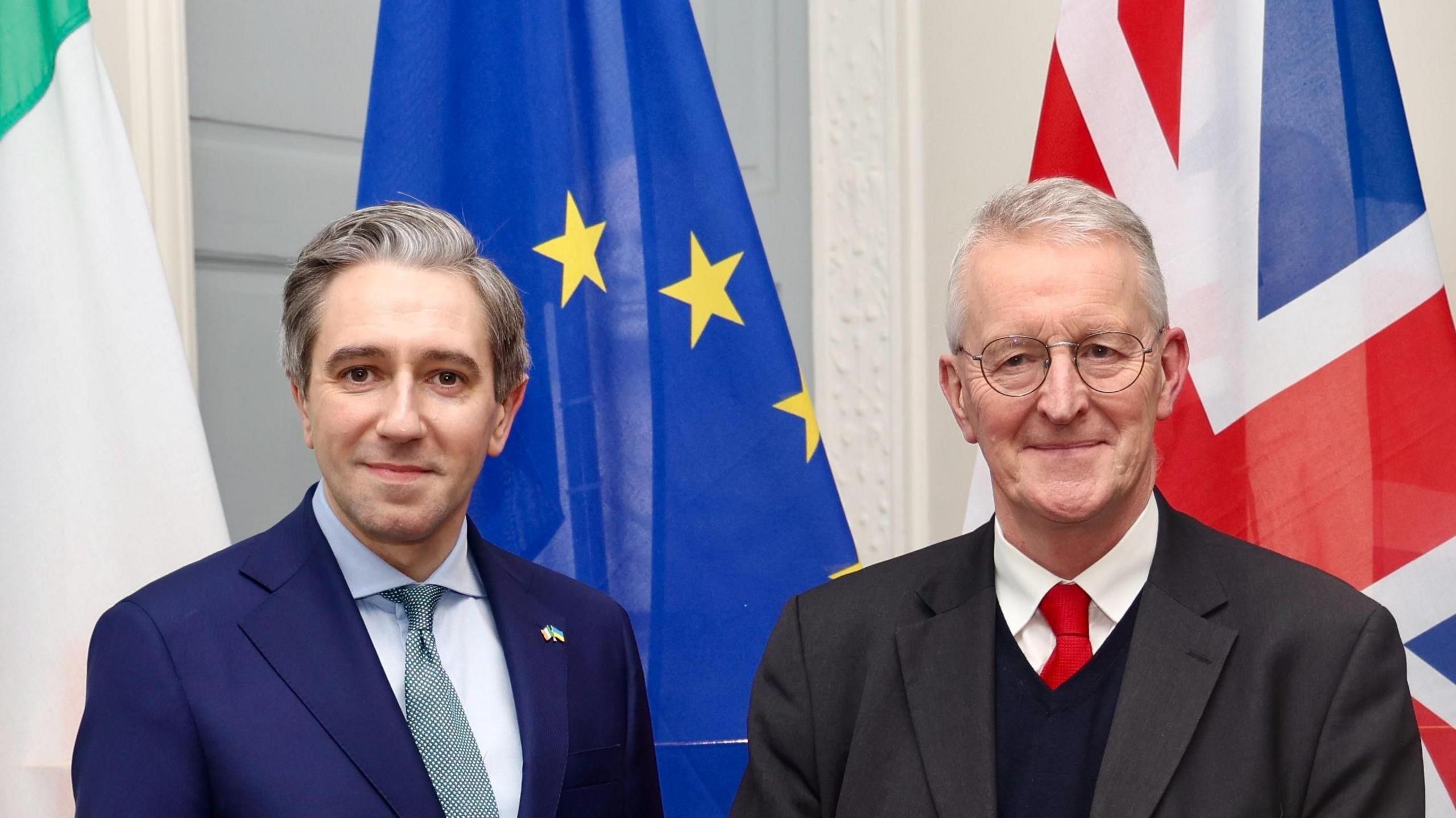 Simon Harris and Hilary Benn stand side-by-side in front of three flags, the flag of Ireland, European Union and Great Britain. Mr Harris has short grey and black hair, and is wearing a navy suit with a baby blue shirt and a patterned tie. Mr Benn has short grey hair, round metal glasses and is wearing a grey suit with a white shirt, red tie and navy V-neck jumper.