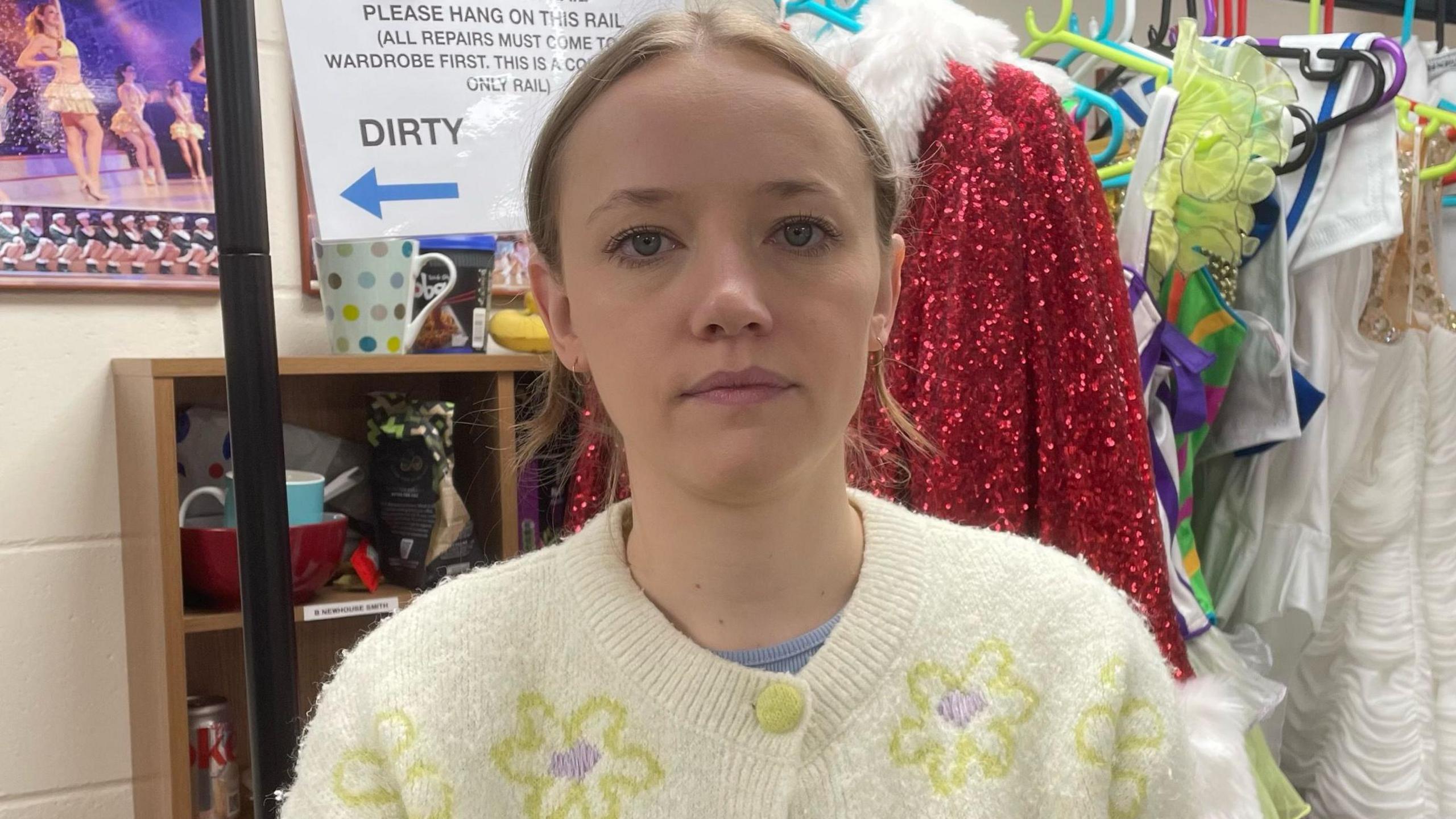 Phoebe Chambers, in a white cardigan which has a lime green button done up at the top. The cardigan has lime green outlines of flowers with a purple circular centre. She is stood in front of a rack of rack of costumes and is looking at the camera. 