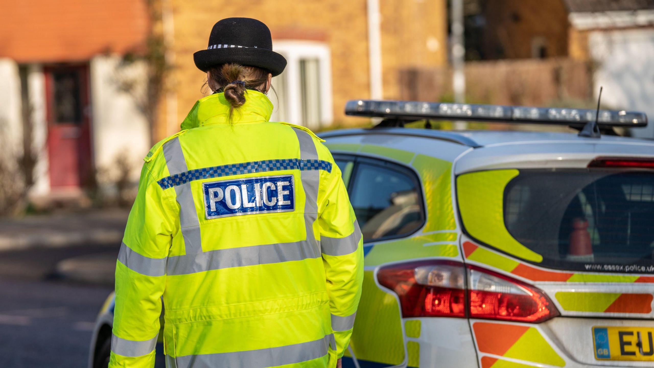 A police officer with their back to the camera wearing a high-vis jacket with police written on it. She is standing next to a police car.