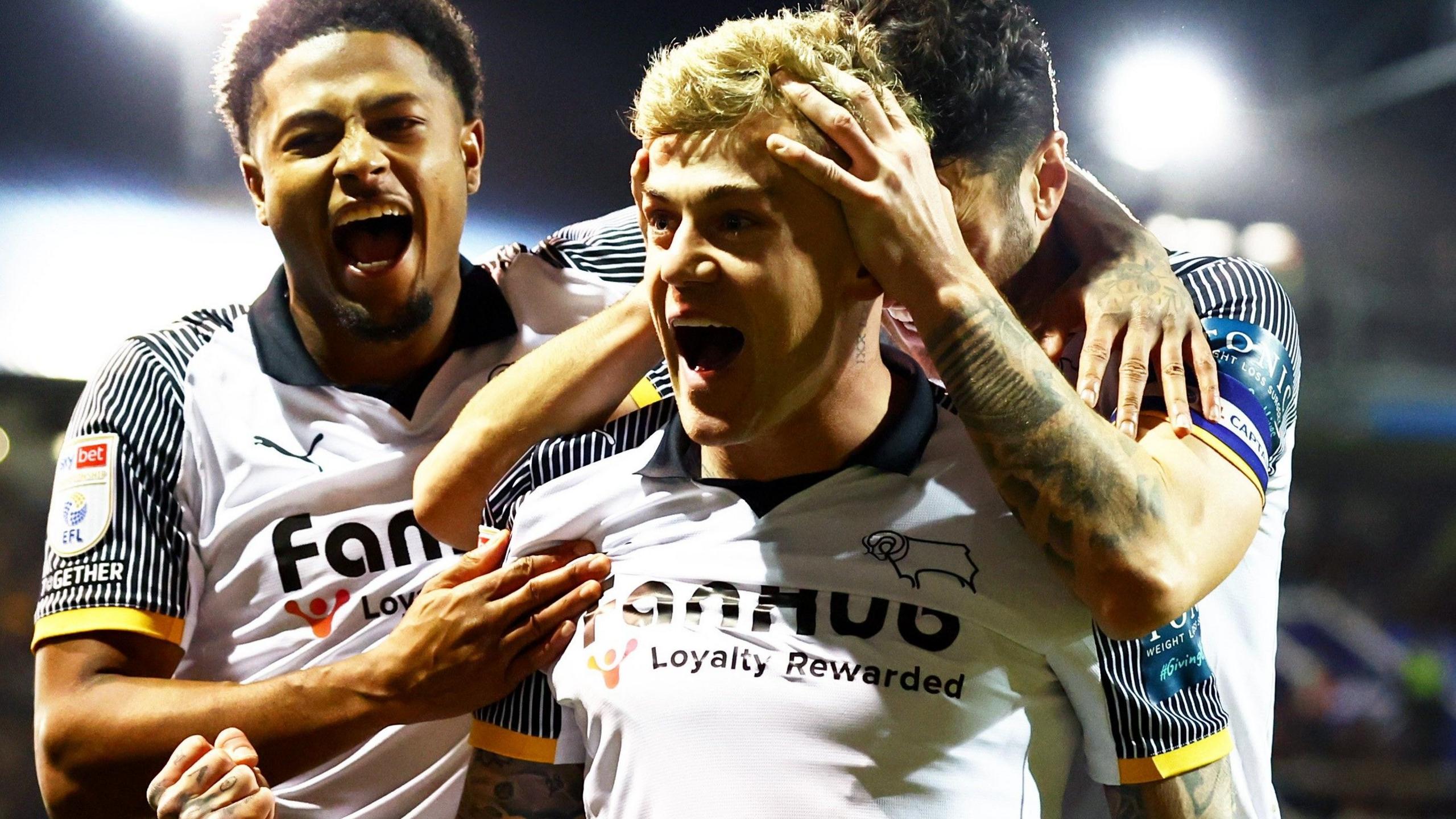 Sammie Szmodics punches his fist towards the fans after scoring the winner at Portsmouth, mouth wide open in celebration with one team-mate's hands on his head and another hugging him and shouting too