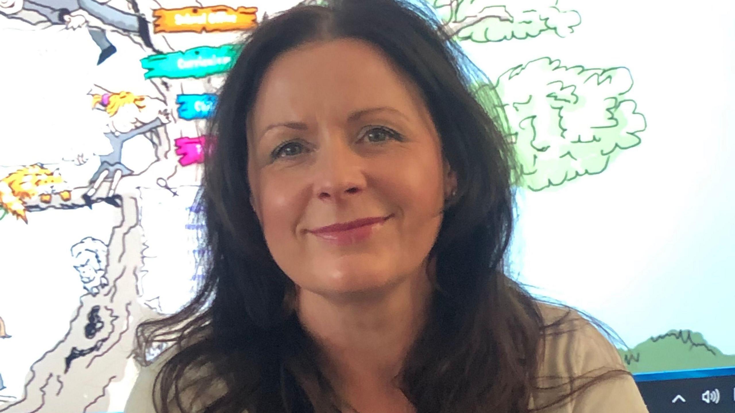 Anne Marie Kenyon has dark brown wavy hair. She sits in front of a whiteboard in her classroom