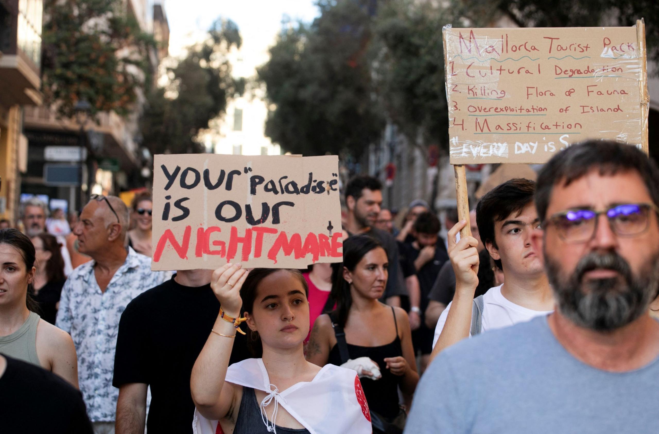 People take part in a protest against mass tourism in Palma de Mallorca, Spain, July 21, 2024. A large group of people walk down a street holding signs. One woman holds a sign that says: "Your 'paradise' is our nightmare"