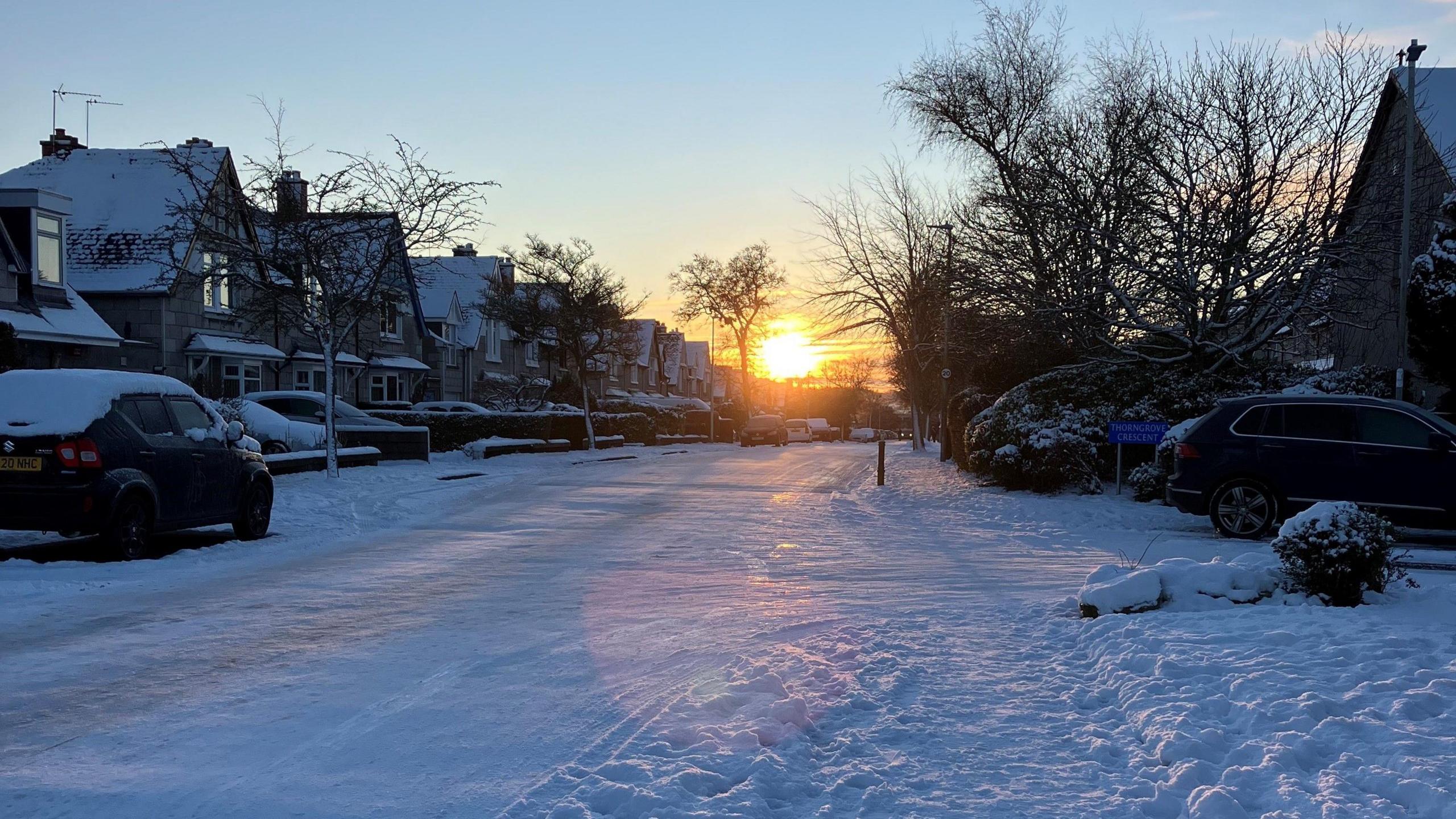 A snowy street in Aberdeen. The road and pavements are covered with snow. Several cars are parked on the street and in drives, which are also covered with snow. The sun is coming up in the background and is orange and yellow against a blue and orange sky.