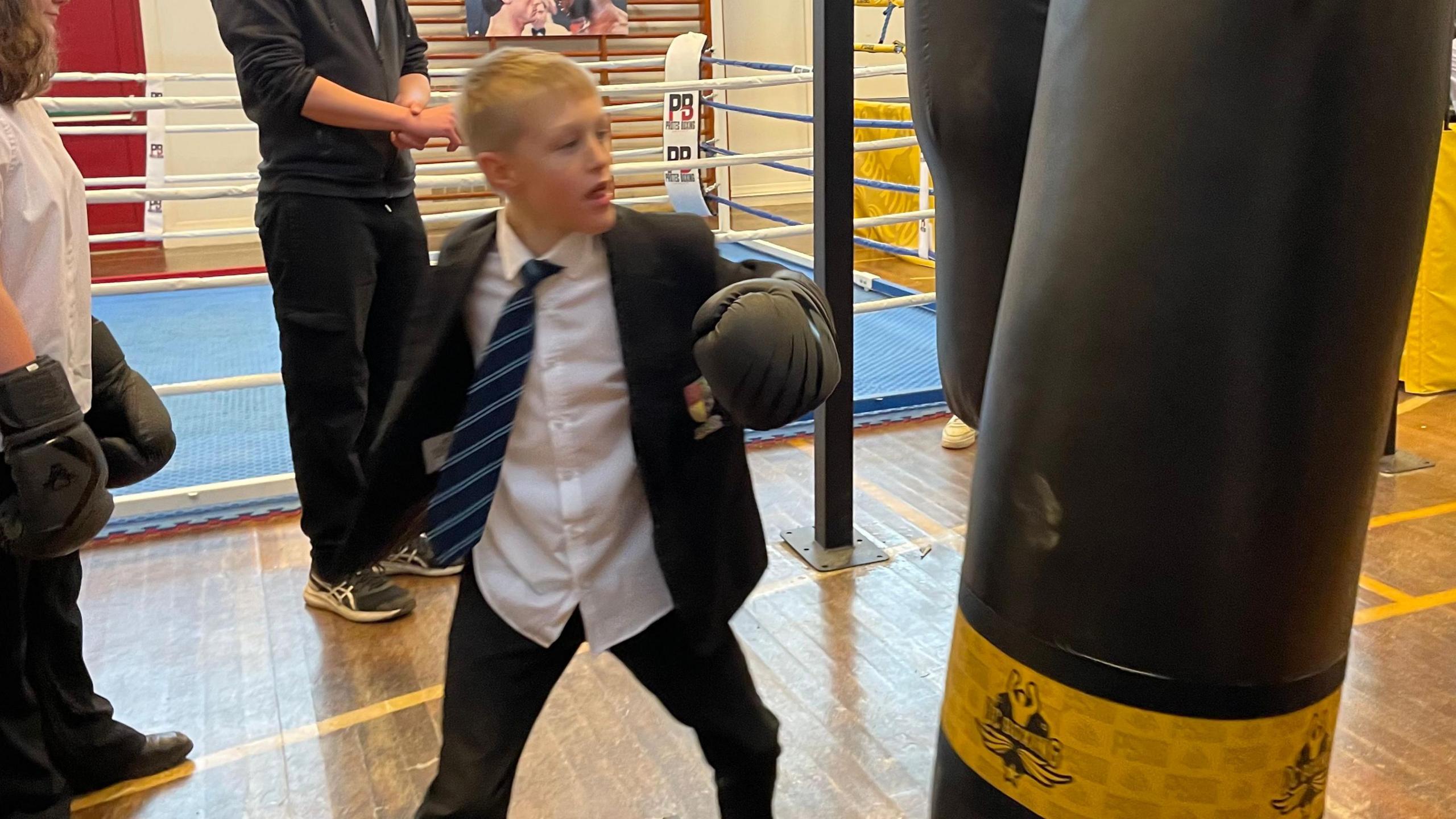 One of the students, in his school uniform, is throwing a punch at a punchbag. Other students are in the background, along with a boxing ring. 