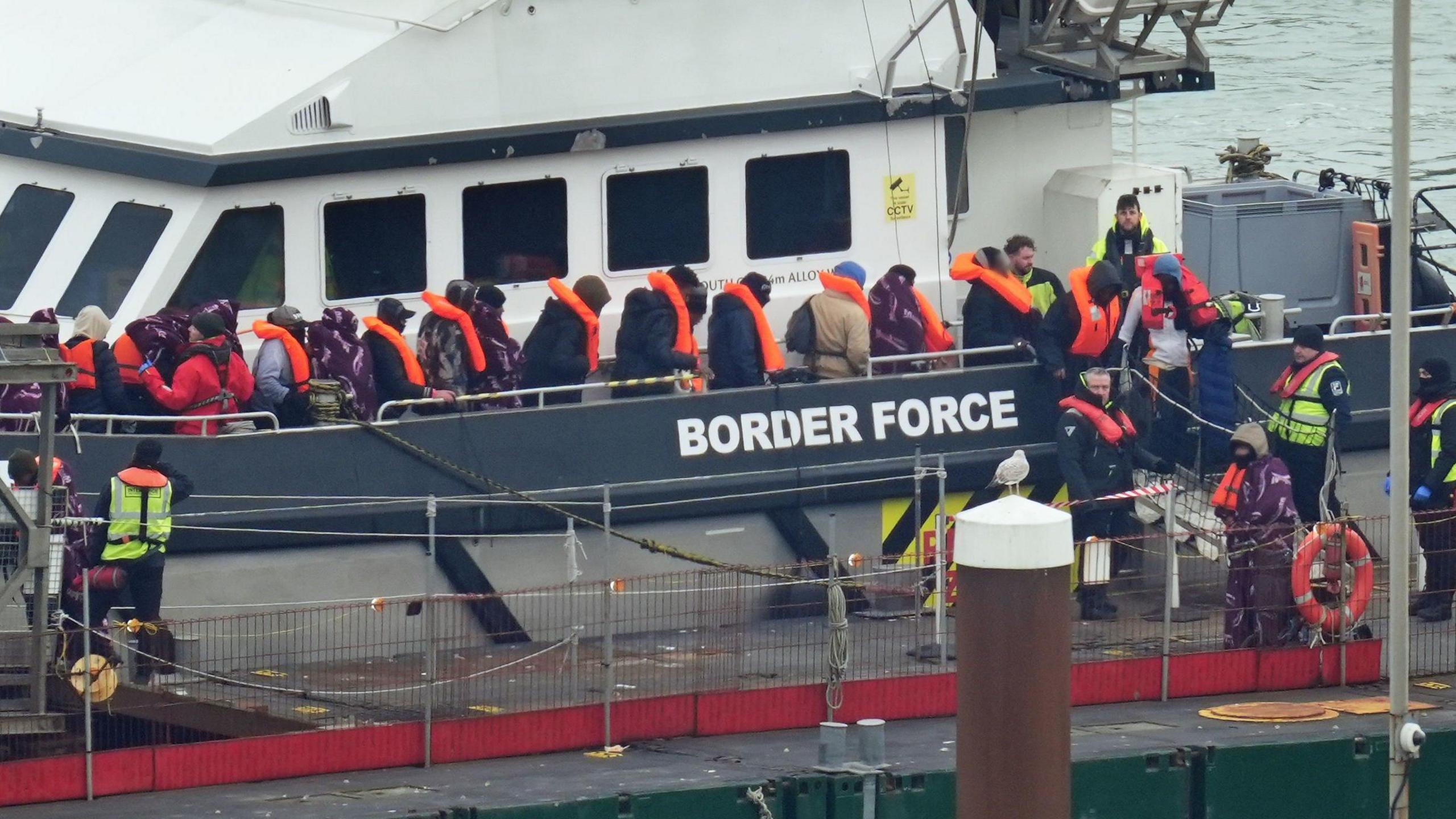 A group of people, thought to be migrants, leave a Border Force vessel. They have orange lifejackets on and look cold. Some are wrapped in blankets and they have hats on. Various staff wear hi-vis jackets around them.