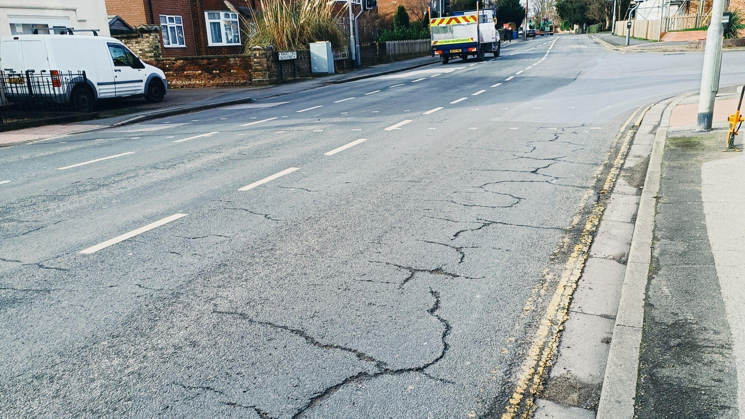 Road with cracks along it. There are houses along the road with a white van in a garden and another van driving along the road.