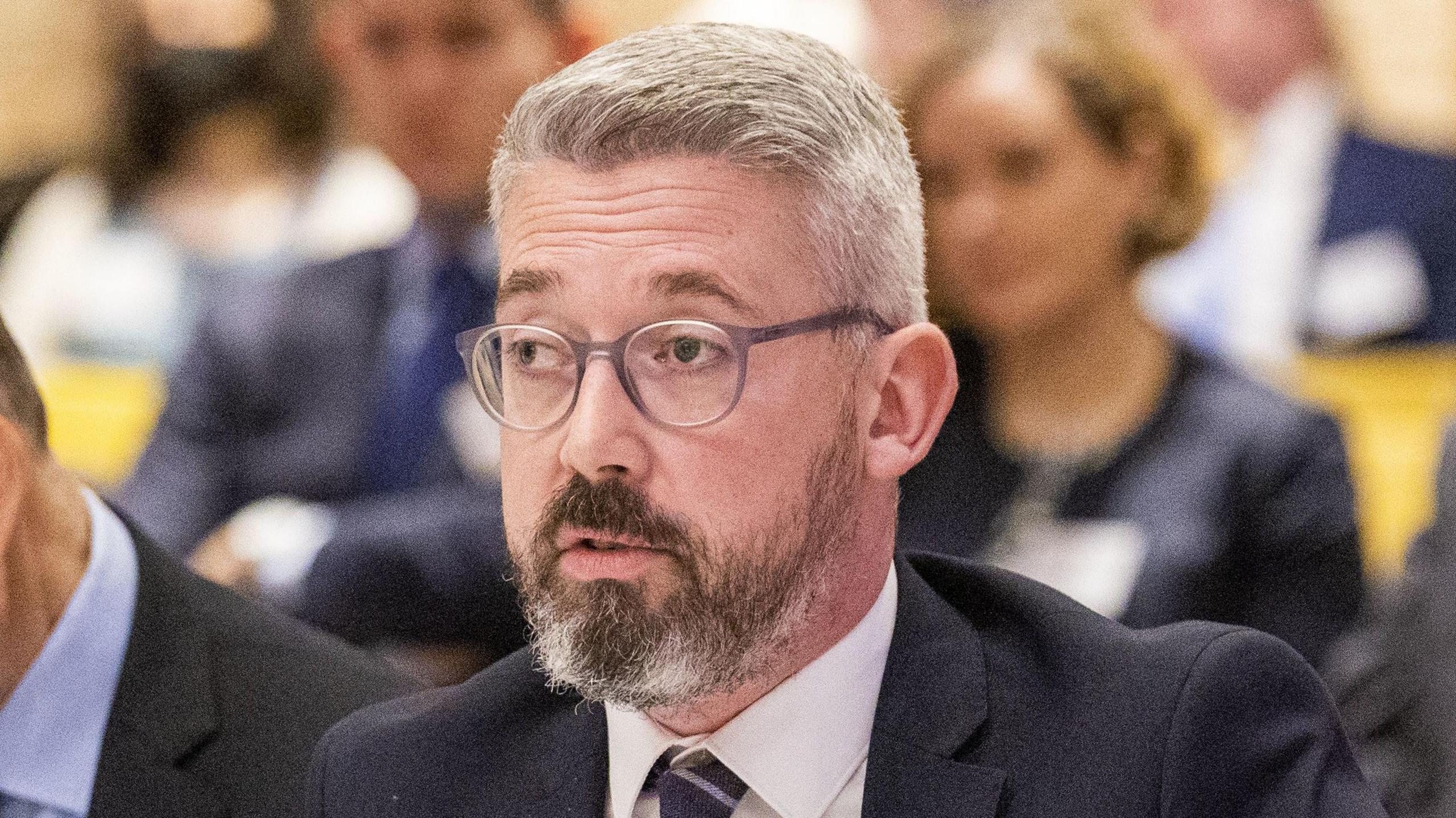 Niall Ó Donnghaile wearing grey glasses, navy suit, white shirt and navy tie.