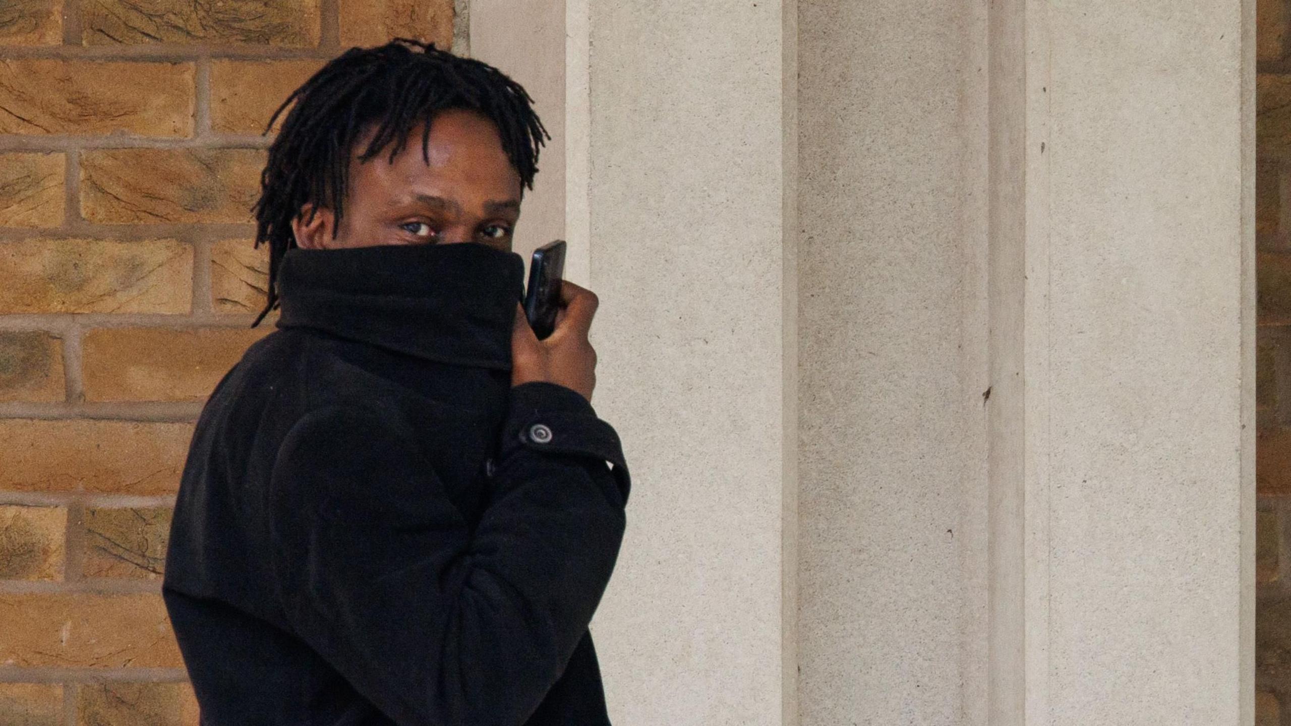 Larry Fraser, wearing a black coat he is pulling over his face to hide from the camera. He carries a phone and looks toward the photographer.