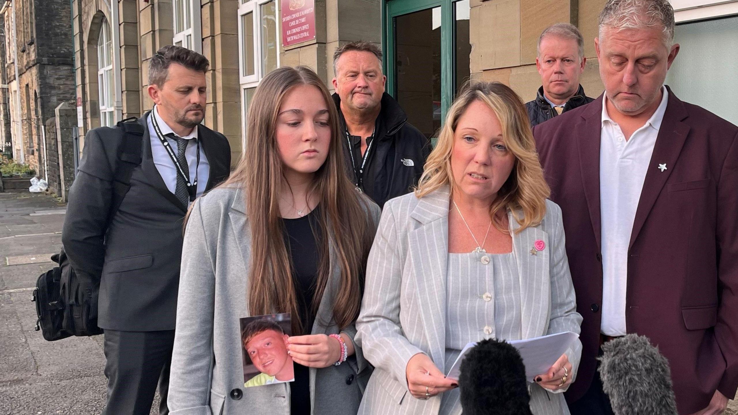 Natalie, wearing a grey pin strip business suit looks up as she speaks from a sheet of paper. A man is next to her in a suit and holding his head down. A young woman is to her right, holding a photograph of Callum in one hand