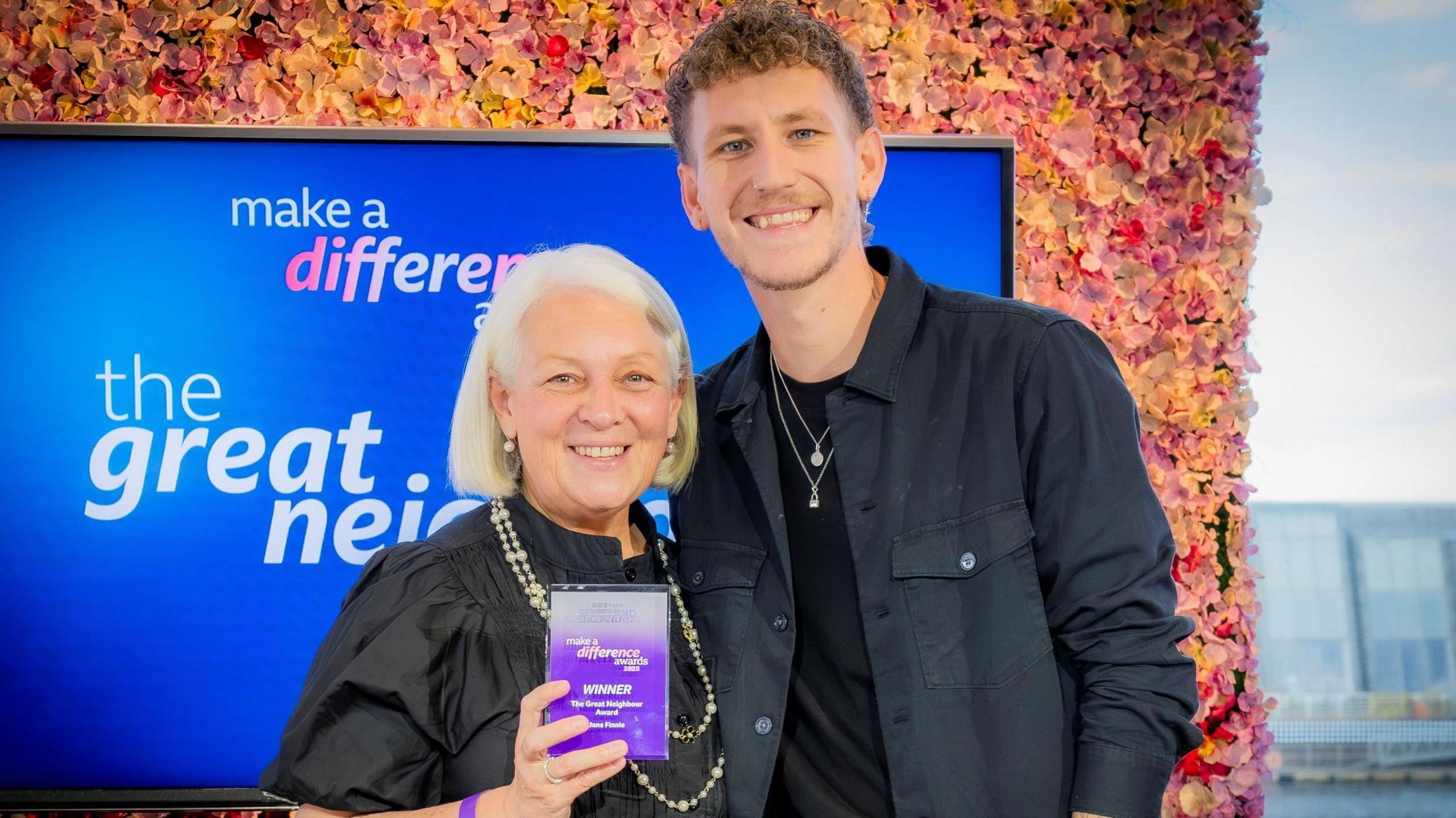 Jacqueline is smiling as she holds the trophy won by her mother Jane. Nathan is standing next to her.
