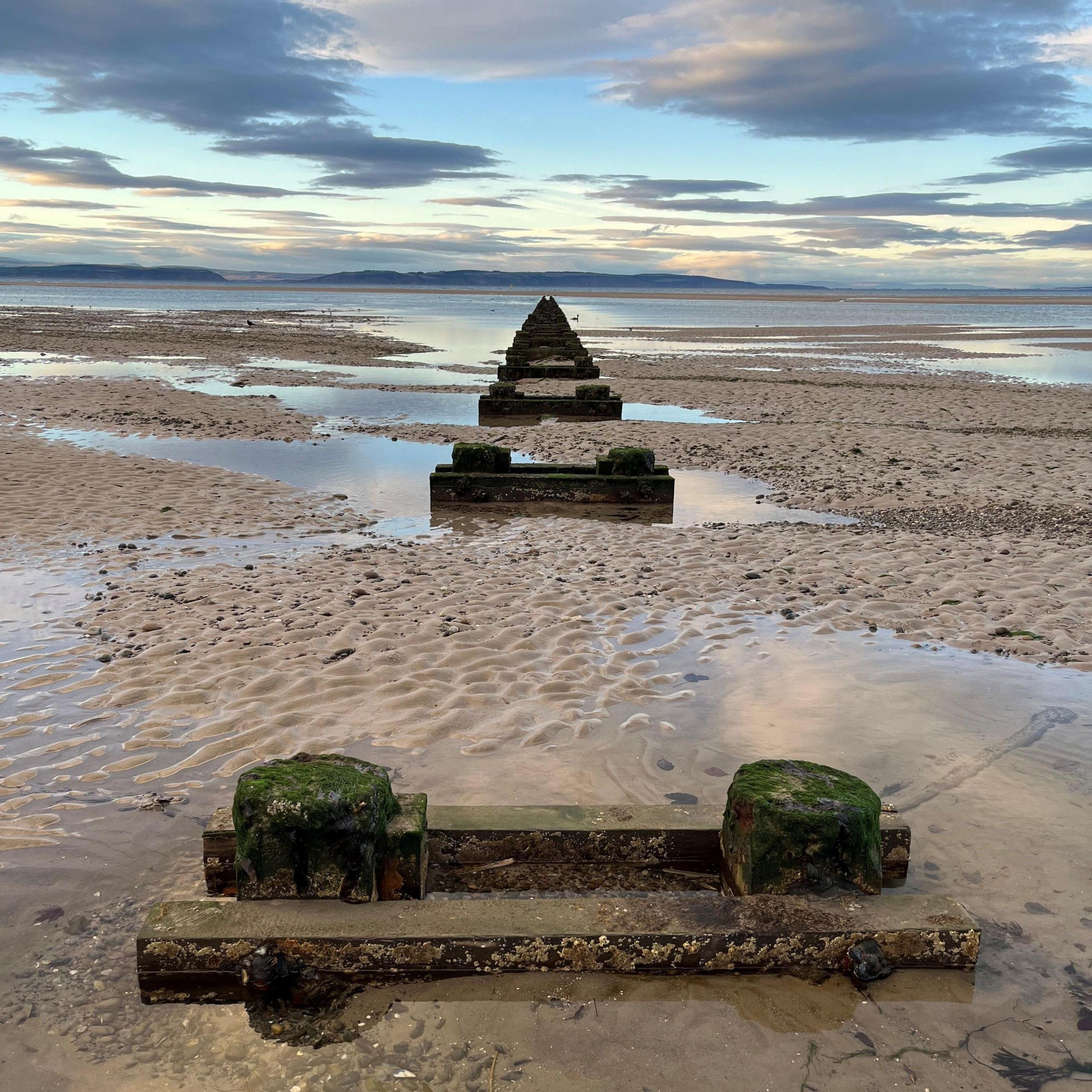 Nairn beach. There is sand on either side of wooden groynes.  Small pools of water stand on the sand. In the distance, there is the sea. Overhead, there is a blue sky with grey clouds.