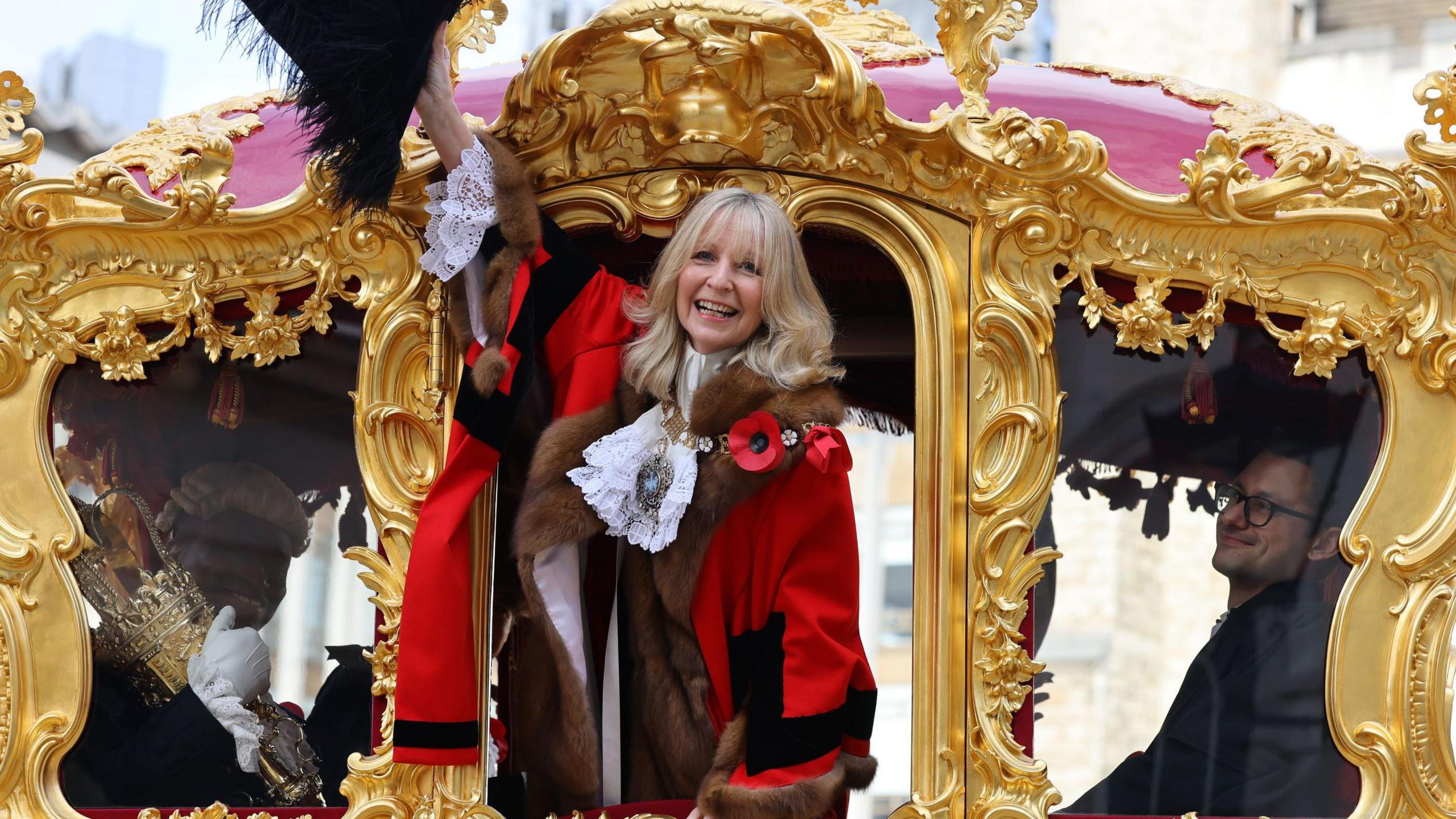 A woman in municipal robes waves from the door of an 18th century golden coach. 