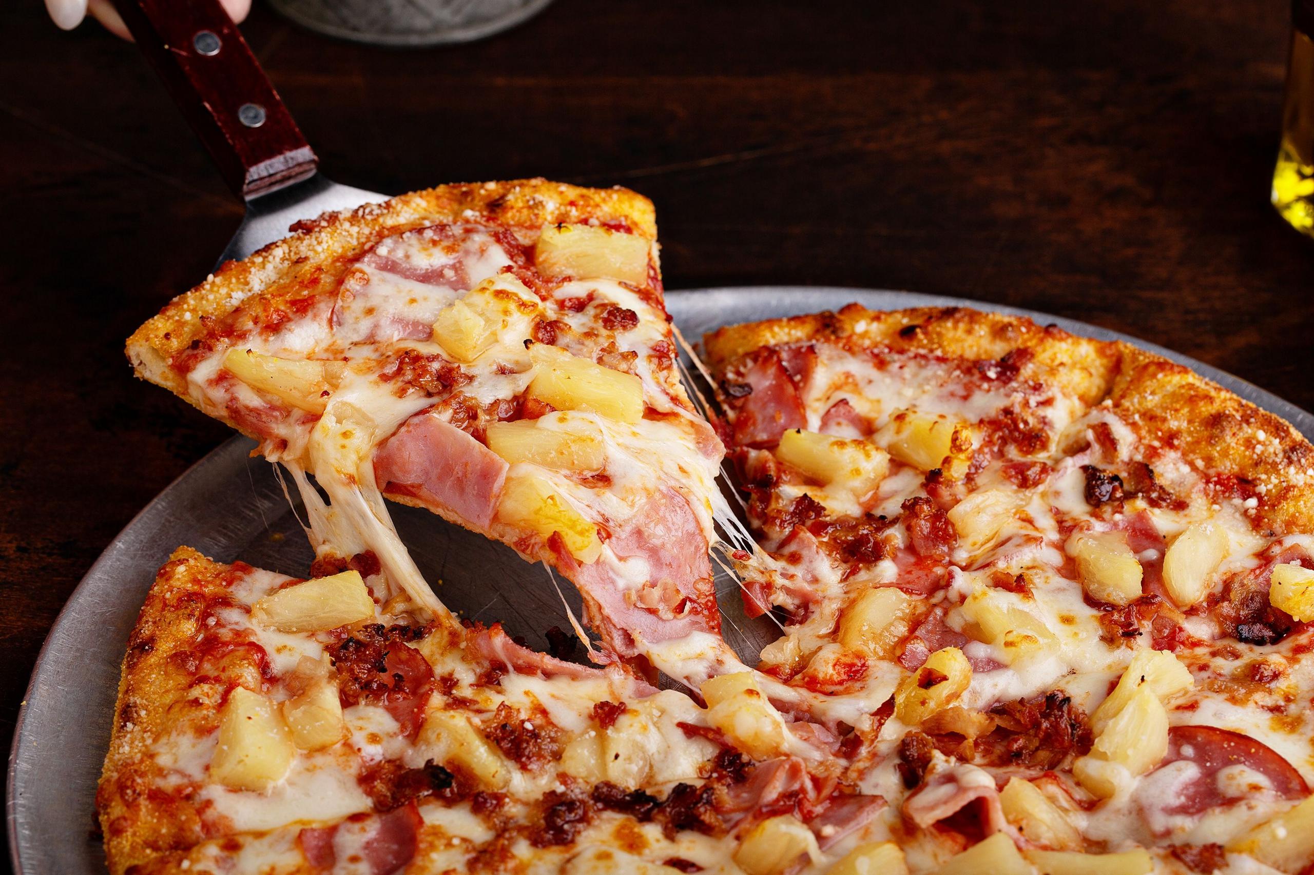 a close up of a pineapple pizza with a pizza slice being lifted up by a knife