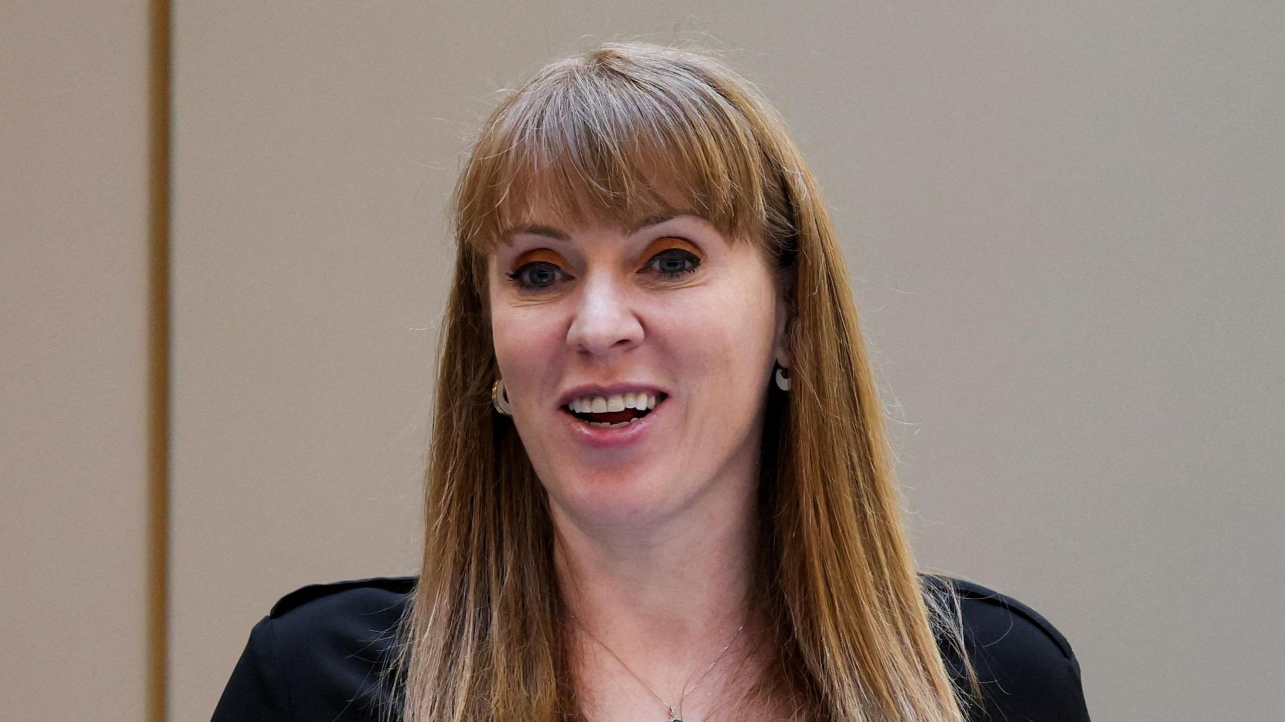 Angela Rayner smiles while looking beyond the camera. She has long red hair with a fringe. She wears a black top as well as earrings and a necklace.