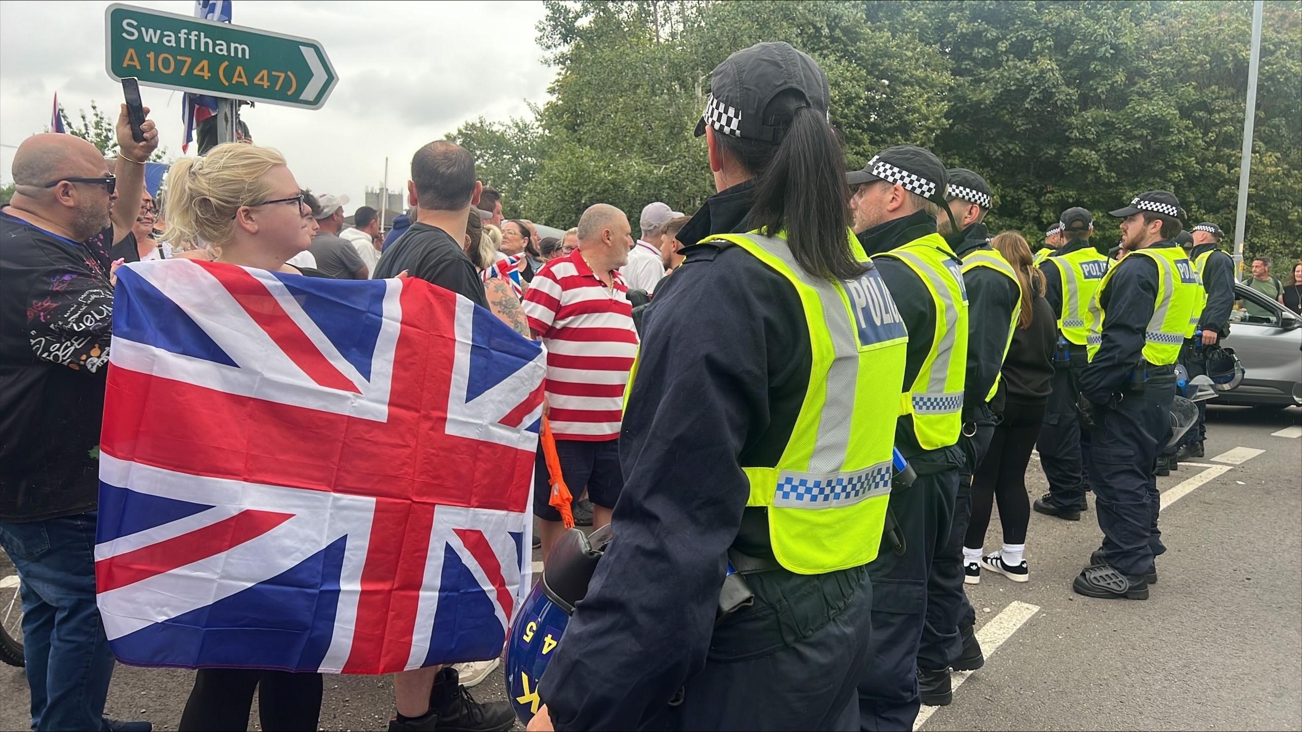 A row of seven police officers stand in a road facing a group of protesters, with trees and a road sign seen behind them.
