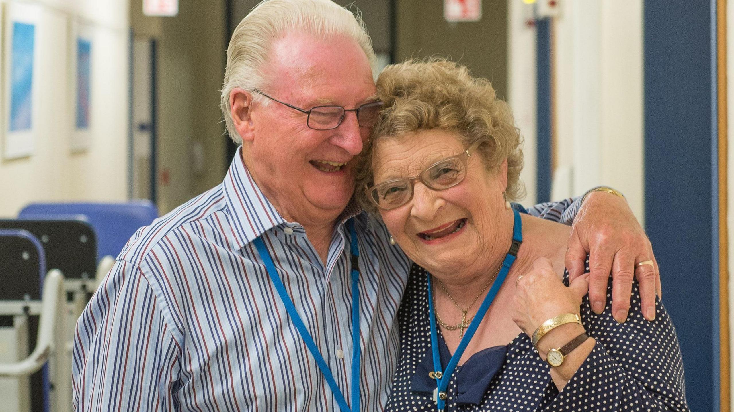 Terry Seston, a man with grey hair and glasses is wearing a blue shirt, with navy and red stripes. He has his arm around his wife Babs, who has curly blonde hair and glasses and is wearing a blue polka dot top. Both are smiling, and Babs is holding Terry's hand, which lies on her shoulder. Behind them can be seen a hospital corridor, with blue wheelchairs along the walls to their left hand side.