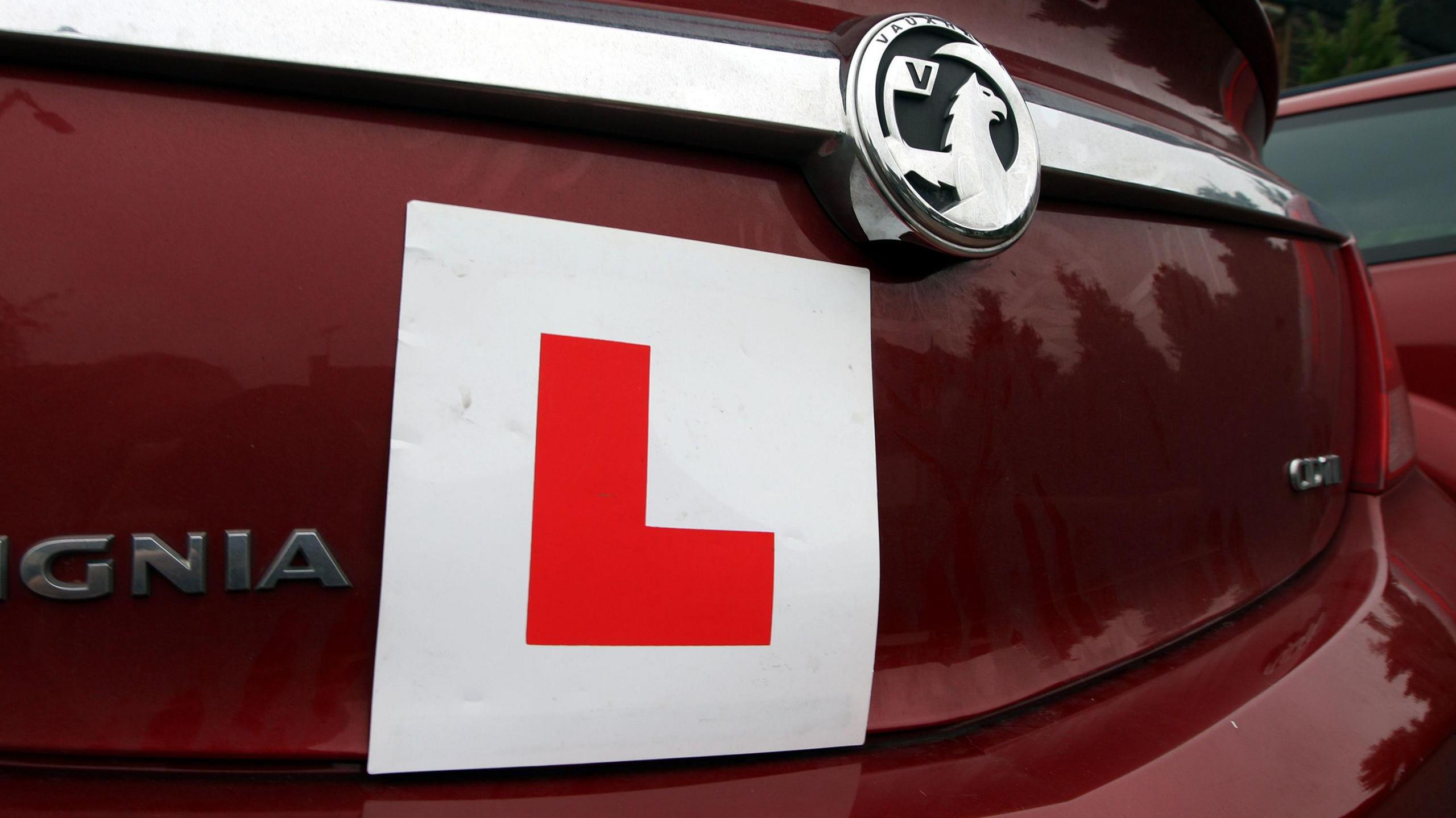 A close-up shows the back of a maroon car, which has a square "L plate" stuck on the rear of the vehicle.
