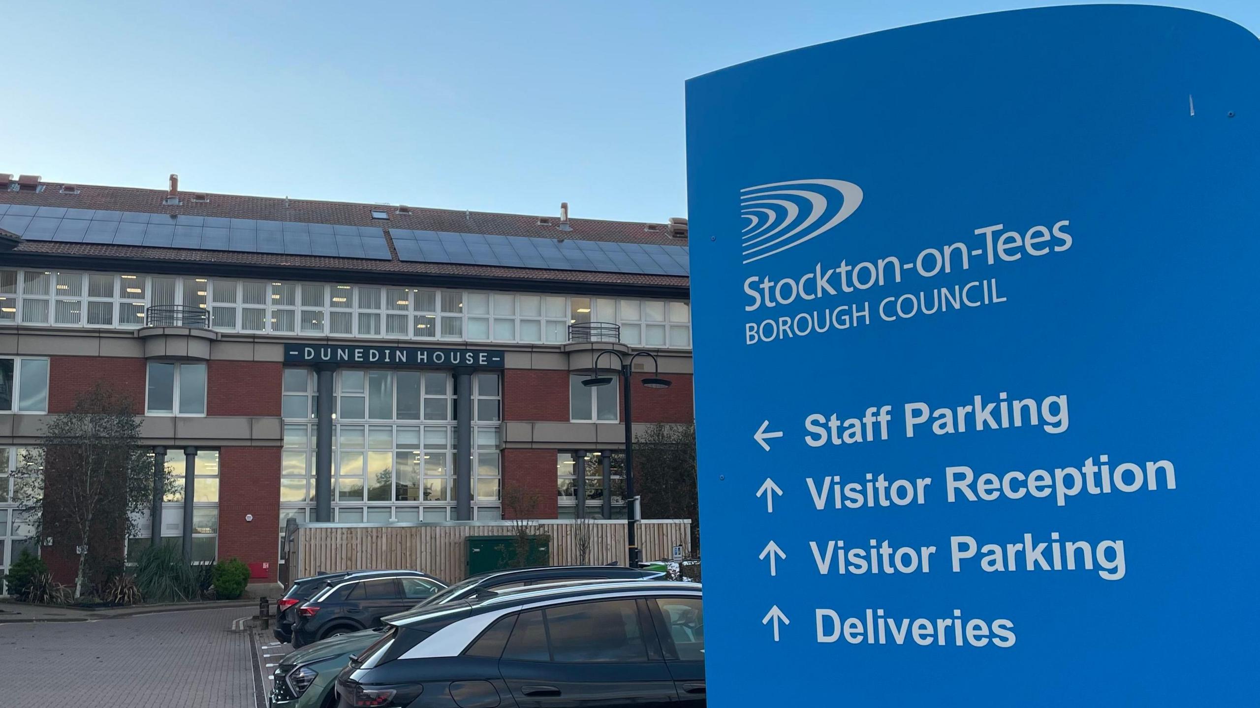 At the right-hand side of the photo is a blue sign outside the Stockton-on-Tees Borough Council building. On it are directions for 'Staff Parking', 'Visitor Reception', 'Visitor Parking' and 'Deliveries'. In the background, part of the car park is visible, and so is the 3-storey building. A sign is at the front of the building that says 'Dunedin House'. The building is made up on predominantly red brick, with many windows and also solar panels on the slanted roof.