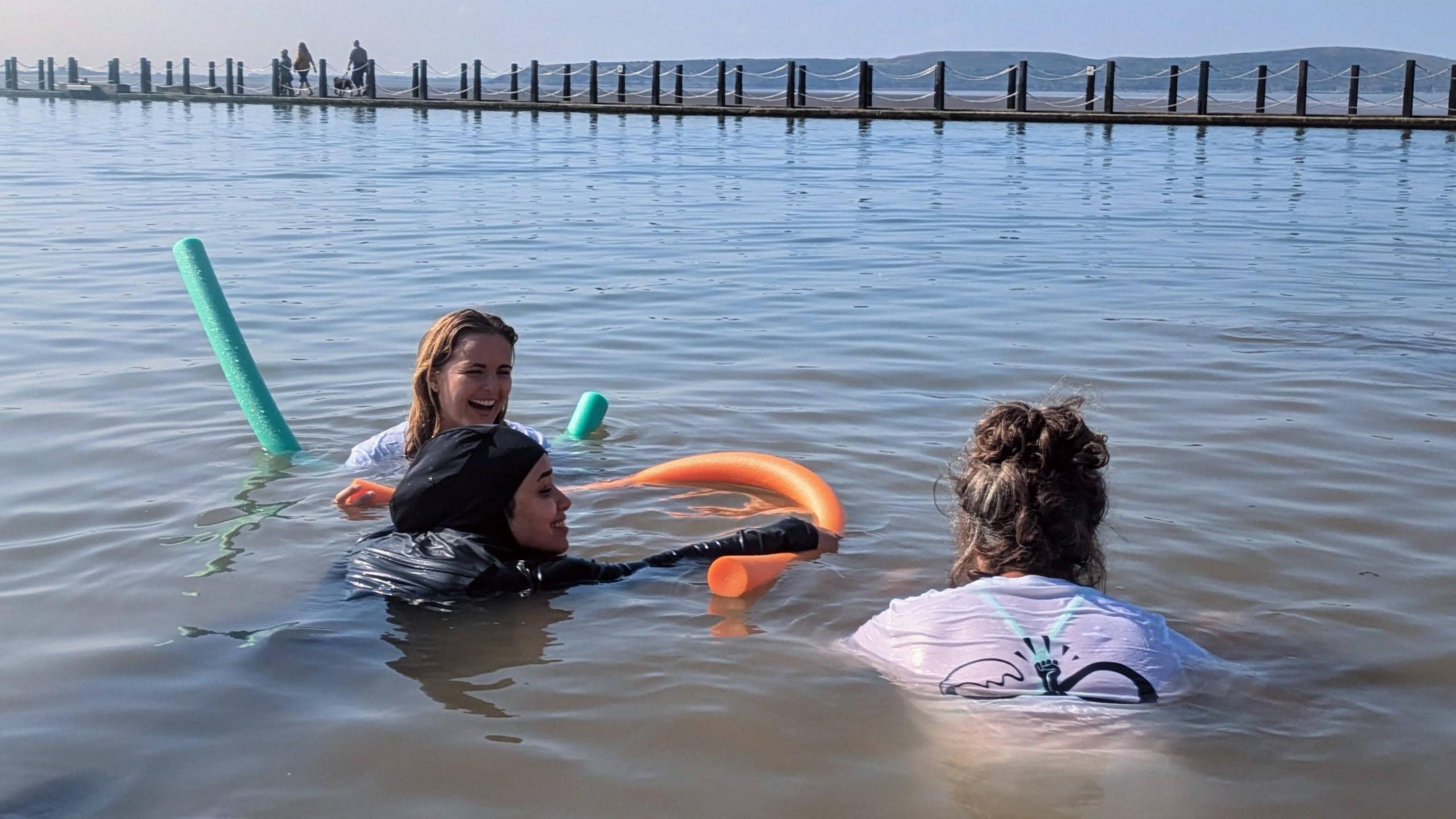 A smiling woman wearing a hijab and black wetsuit, lying on top of an orange pool noodle in Weston-super-Mare's Marine Lake. There are two women wearing white rash vests laughing alongside her.