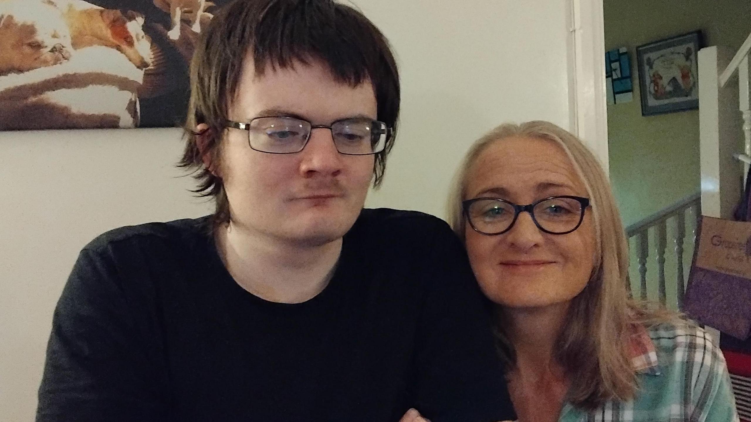 Nicola Kilcoyne is sitting beside her son Jack in a living room. Jack has dark brown hair and is wearing a black t-shirt and glasses. Nicola is wearing a teal plaid shirt and black glasses, with shoulder-length light hair. There is a picture on the wall behind them.