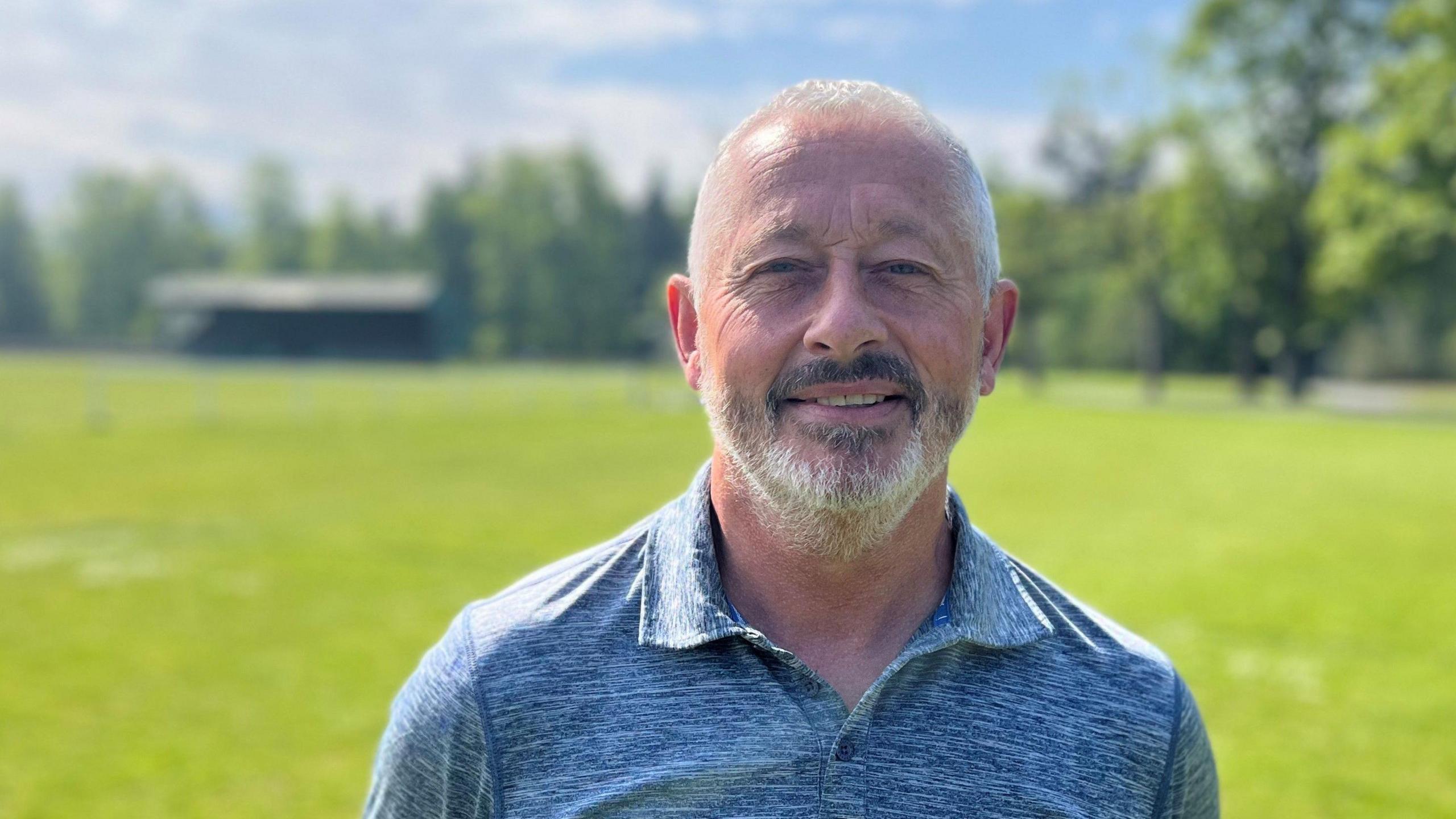 A man with grey hair and grey beard, wearing a blue polo shirt, looks at the camera. A football pitch and small stand are in the background