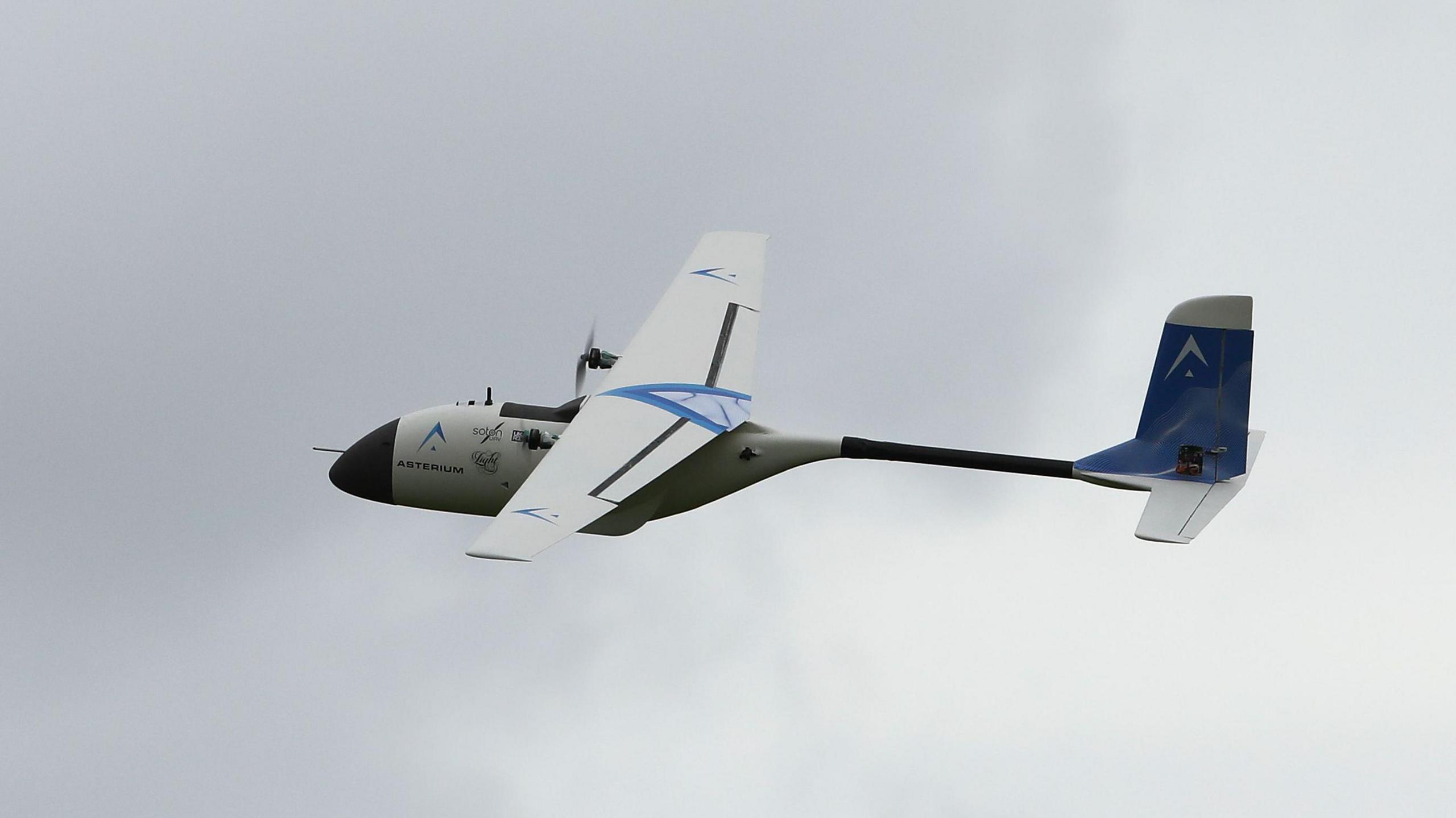 A blue and white drone in flight in a cloudy sky.