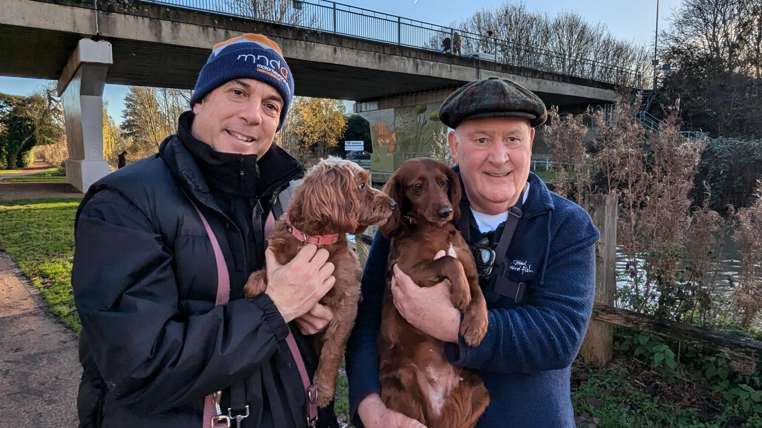 Two men wearing warm clothes and hats smile for the camera as they each hold a brown dog. They are standing on a footpath on a sunny day with a canal and bridge in the background.