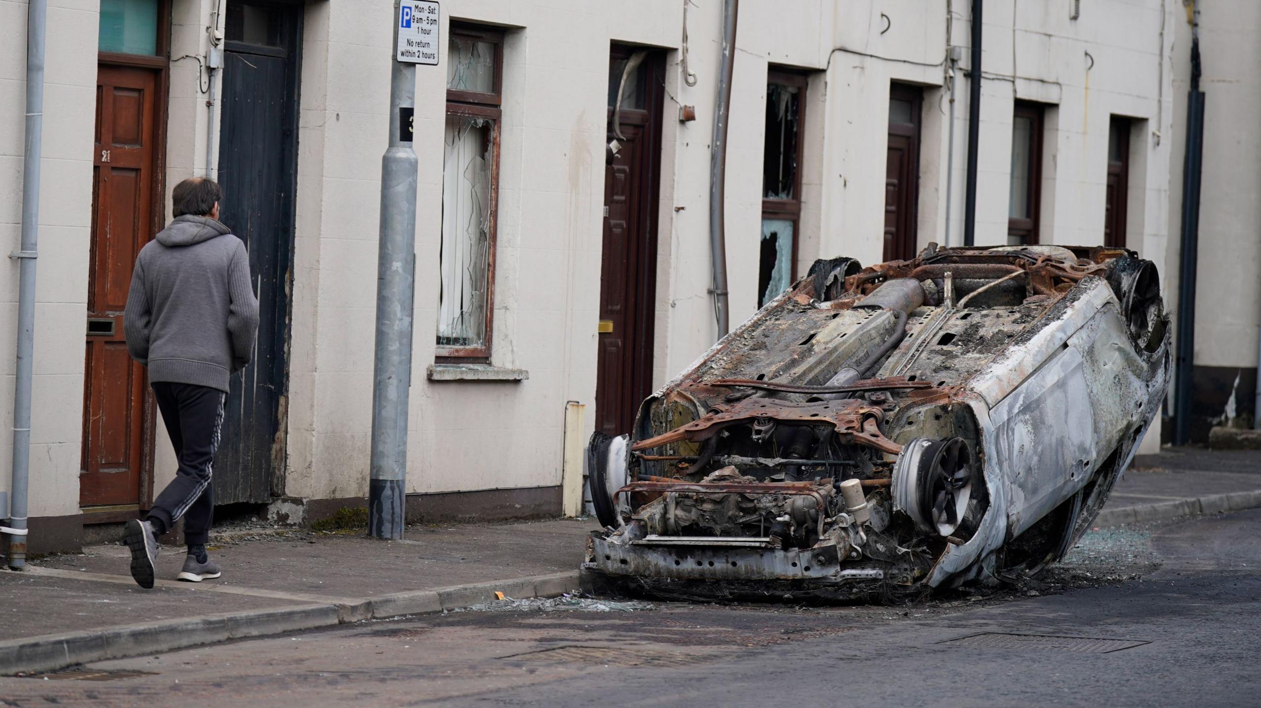 A man wearing a grey hoodie and tracksuit bottoms walks past an overturned and burnt out car on the street. There are a row of white terrace houses in the distance.