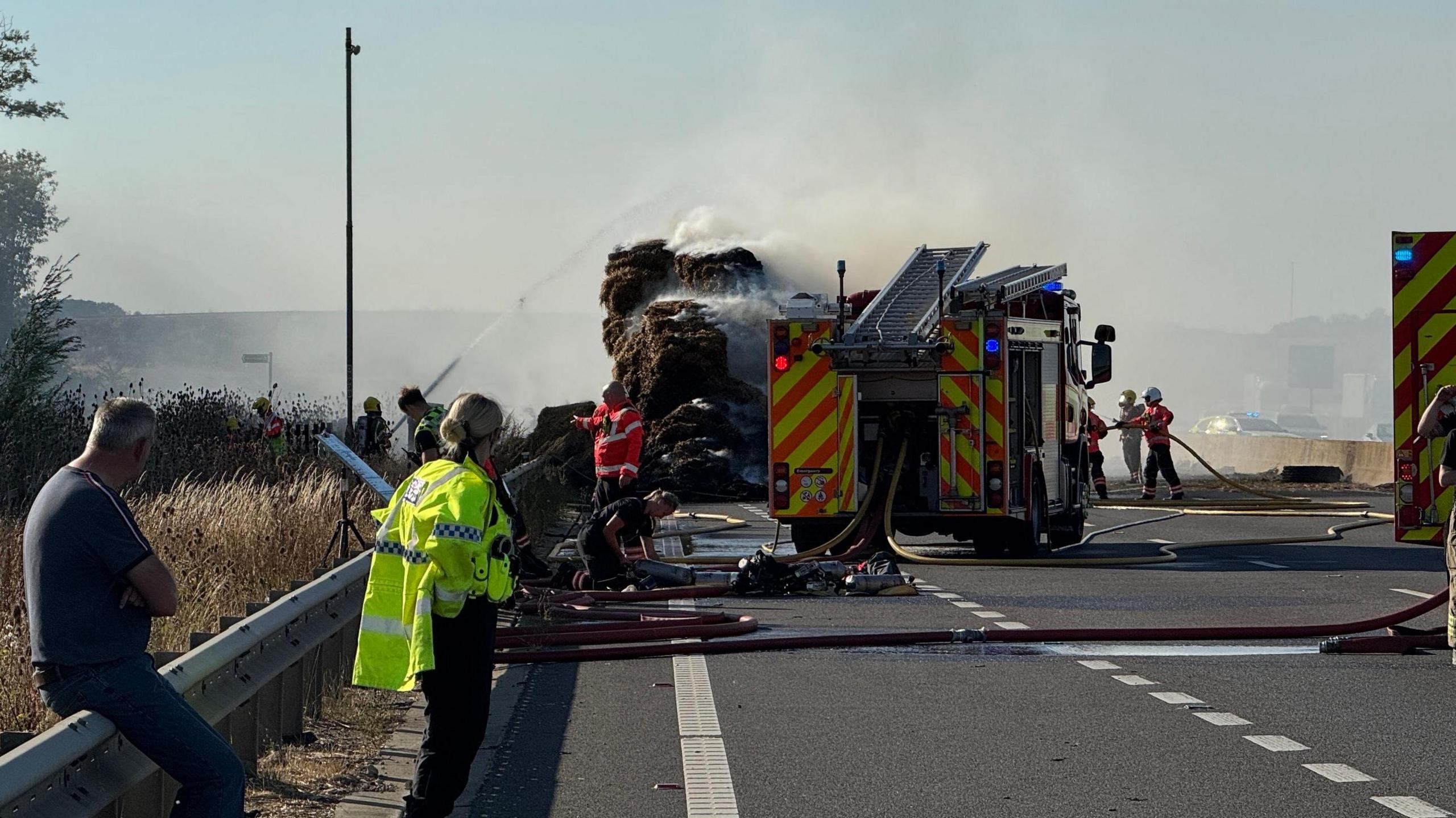 A fire engine on a dual carriageway. Beyond it, a vehicle is on fire. Firefighters can be seen spraying the vehicle with water. There is a man sitting on the crash barrier on the left. A police officer in a hi-vis jacket is standing just in front of him looking down the road at the fire.