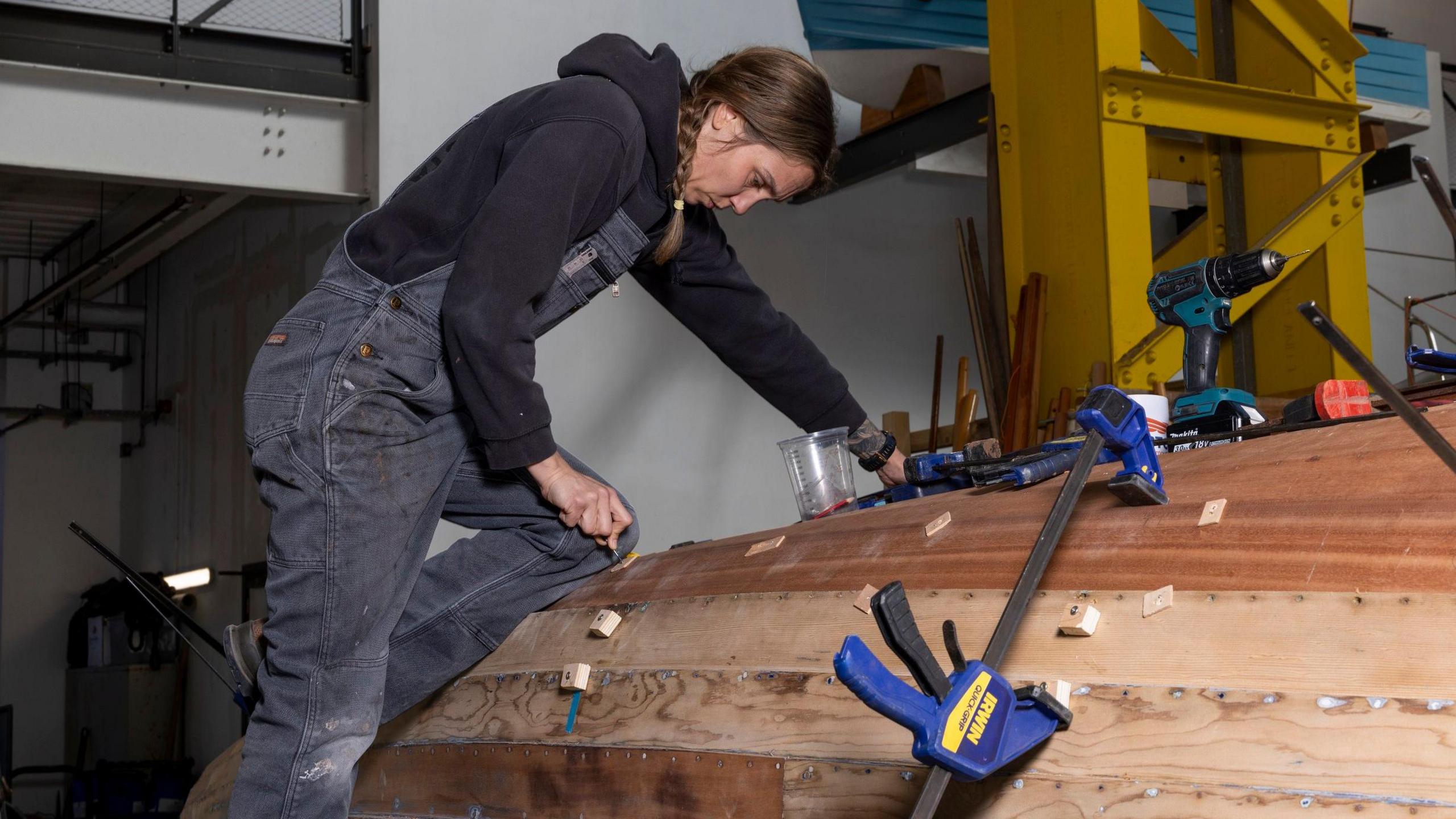 Betzy Shell, wearing dark dungarees, kneels on the upturned hull of a boat inside a workshop