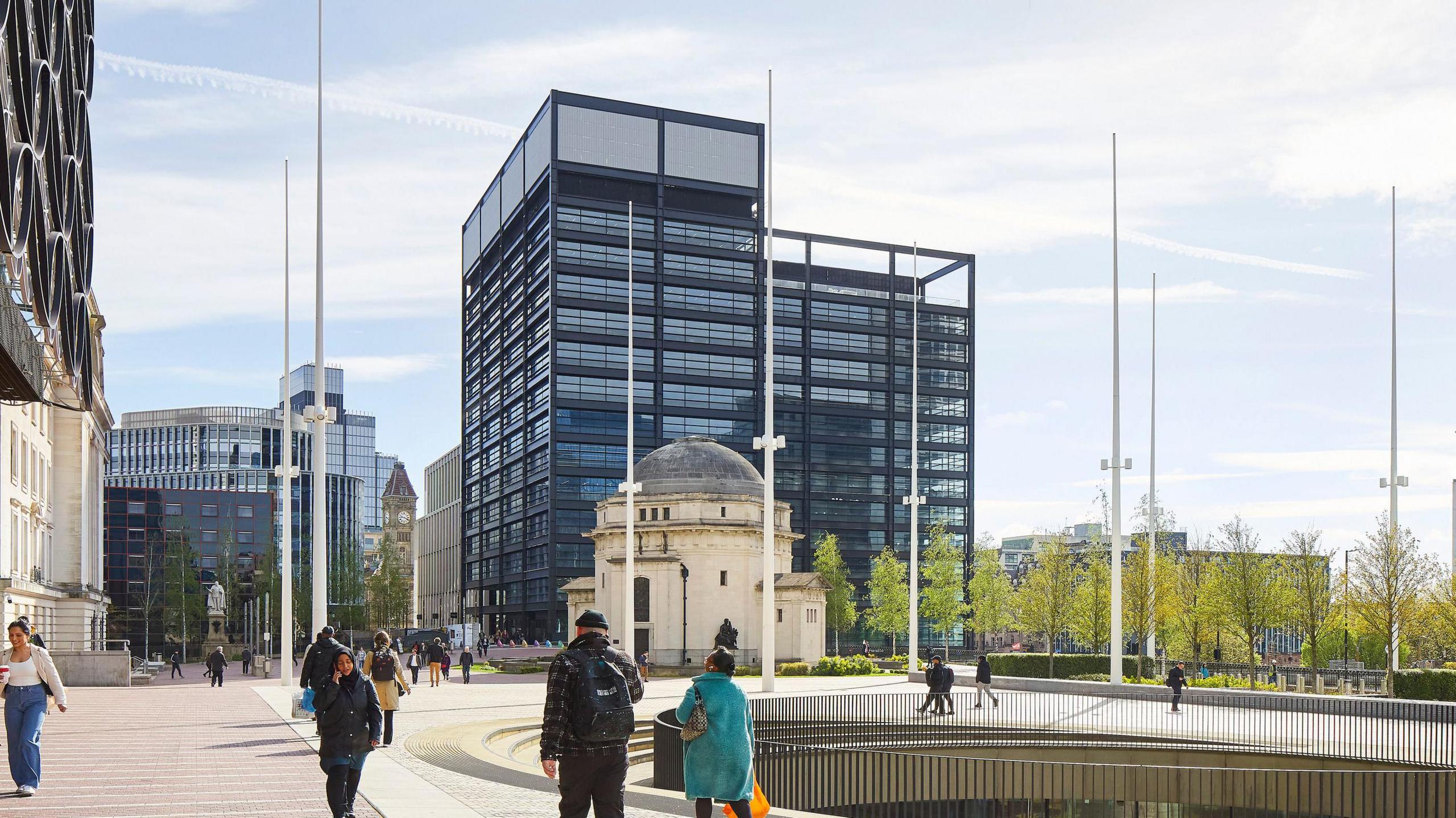 A grey rectangular, high-rise building in the distance, with a small, cream circular building in front. People are walking around the open space in front of the buildings. 