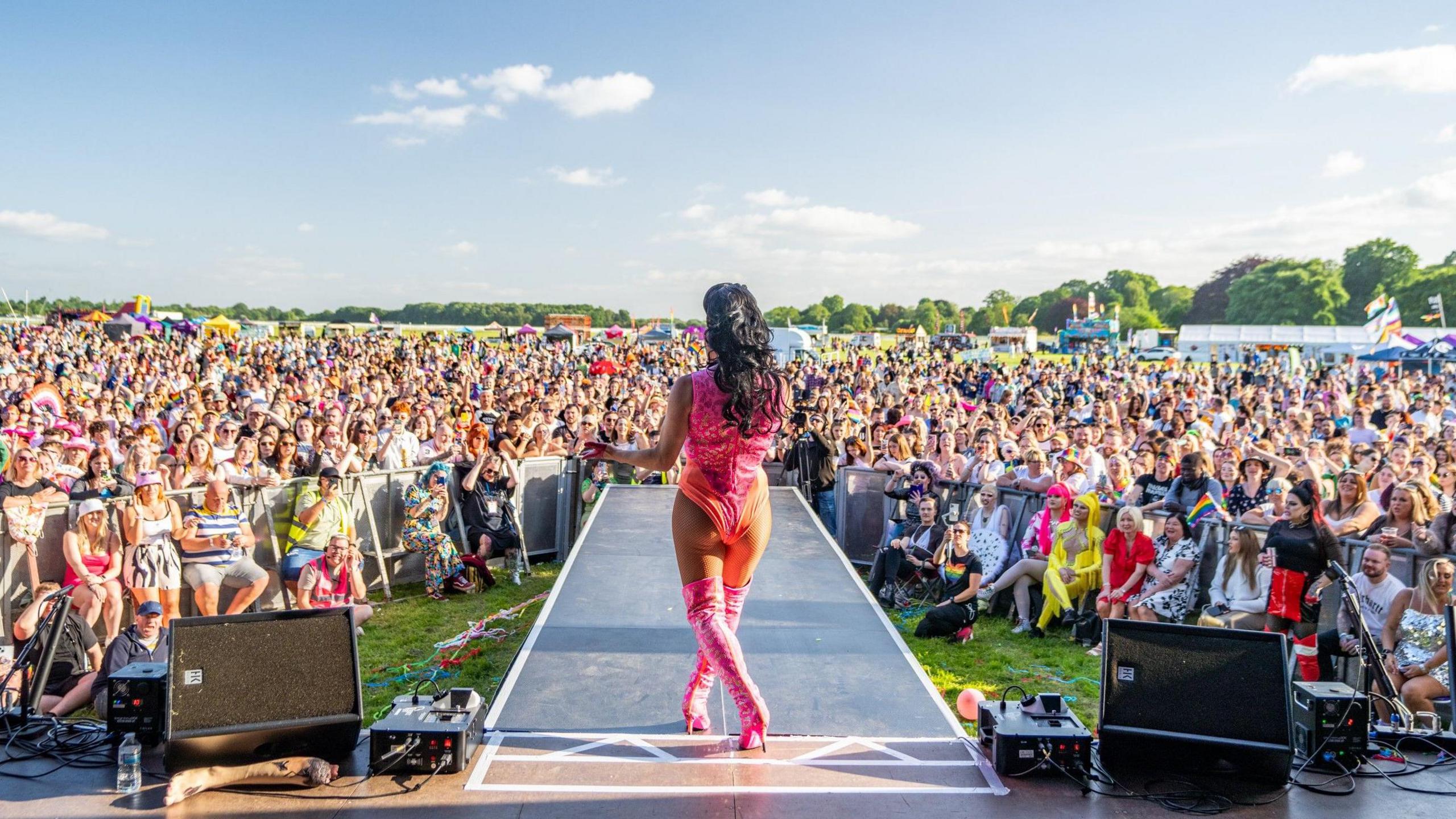 A drag performer in a pink and orange leotard with pink stiletto boots performs in from of a crowd of hundreds of people.
