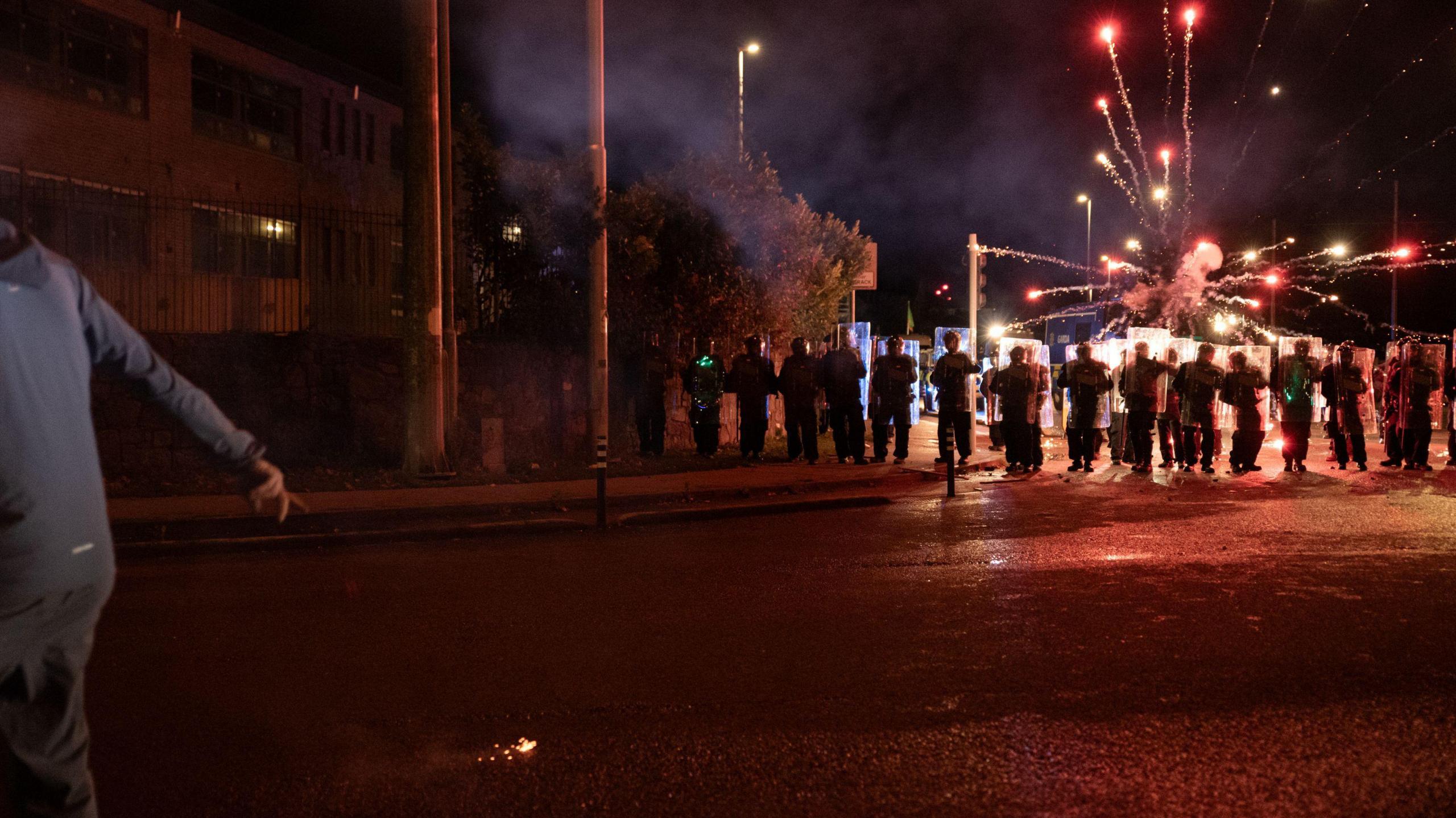 Riot police are lined up across the street. In the sky above them red fire works are visible. There is a person in a grey tracksuit at the front of the photo. The sky is dark with buildings n the background. 