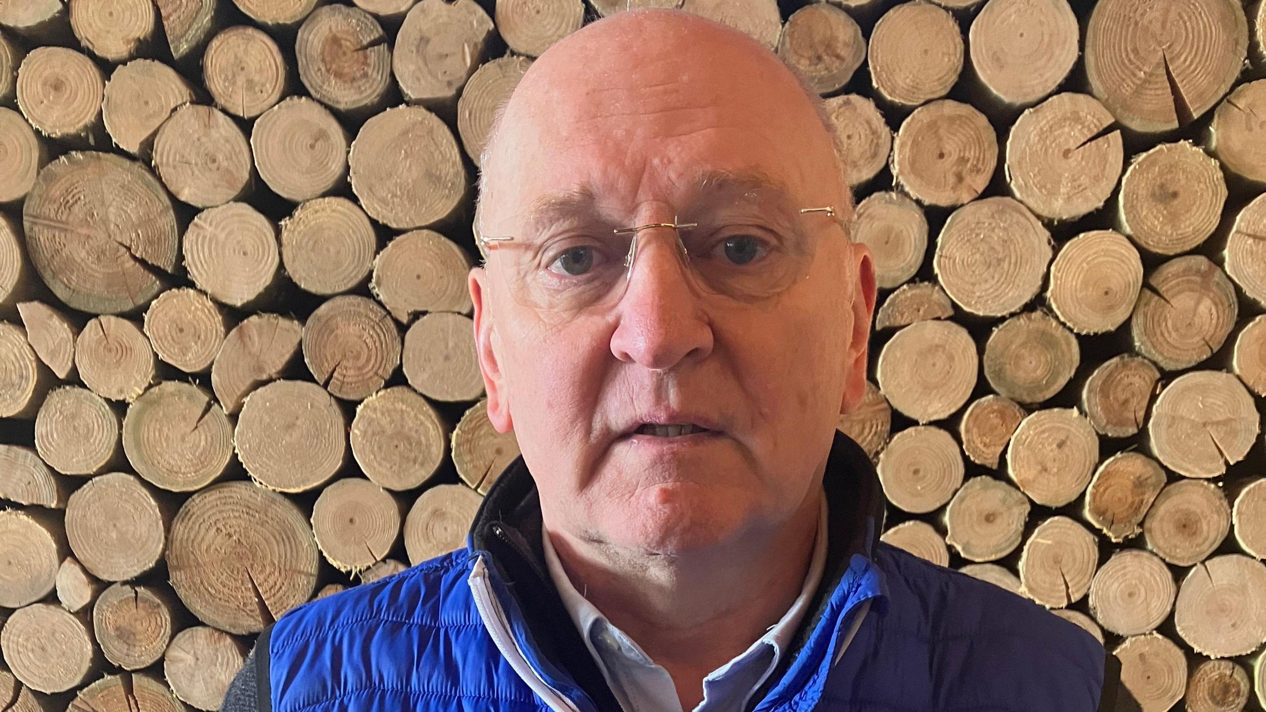 Rowland Rees Evans standing in front of a pile of logs. He is wearing a blue jacket and has glasses on.