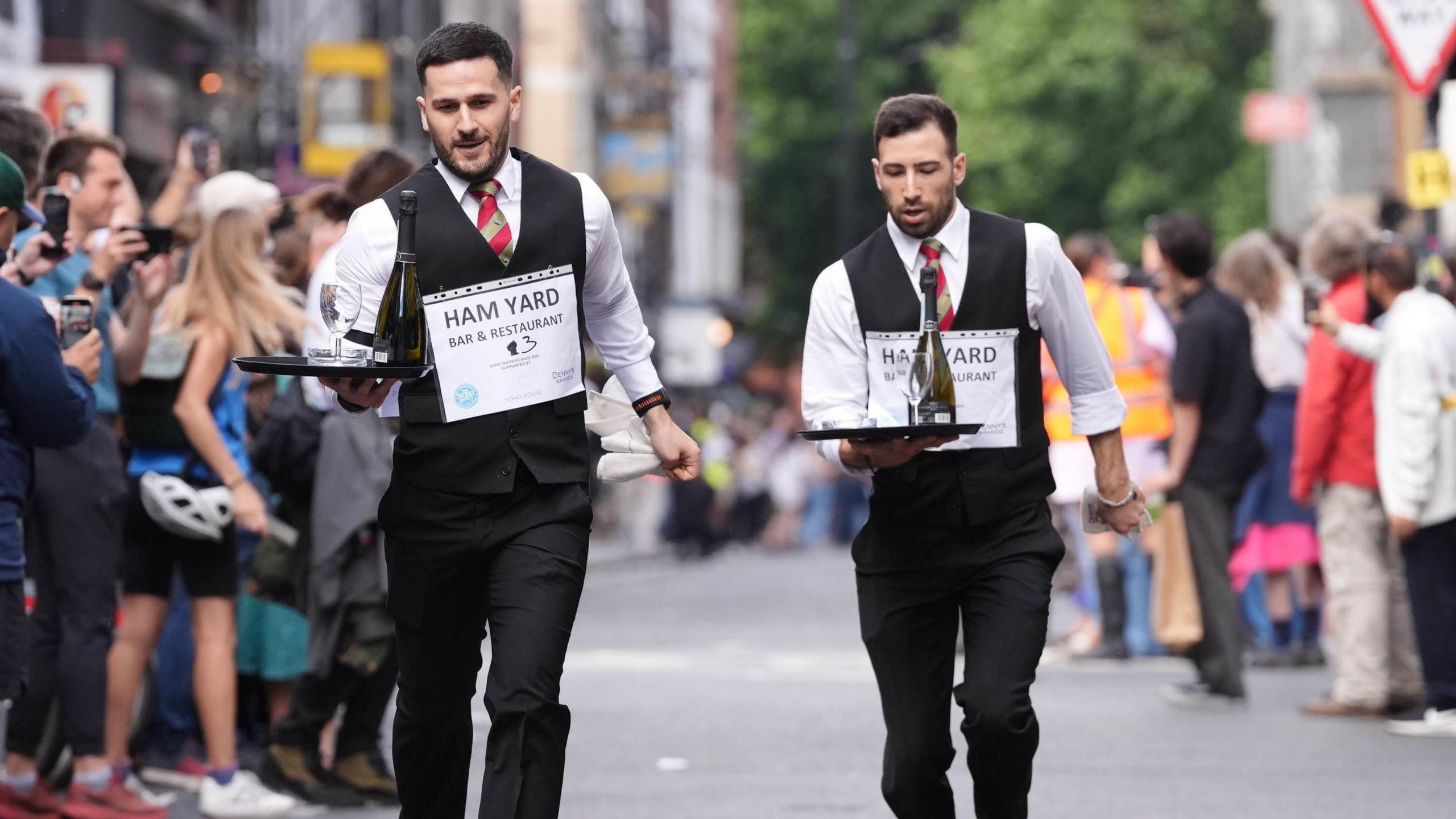 Two young men wearing dark suits and red and grey striped ties, running along a crowd-lined road holding trays with a bottle of wine, glass and ashtray on.
