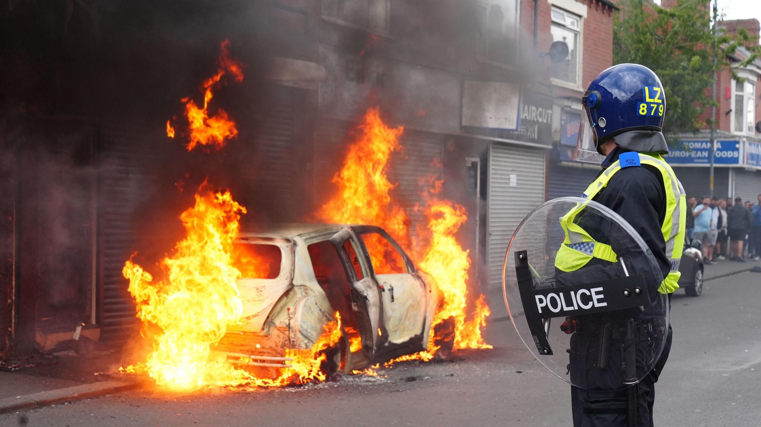 A police officer wearing a helmet and shield looks at a white car on fire outside a shop.