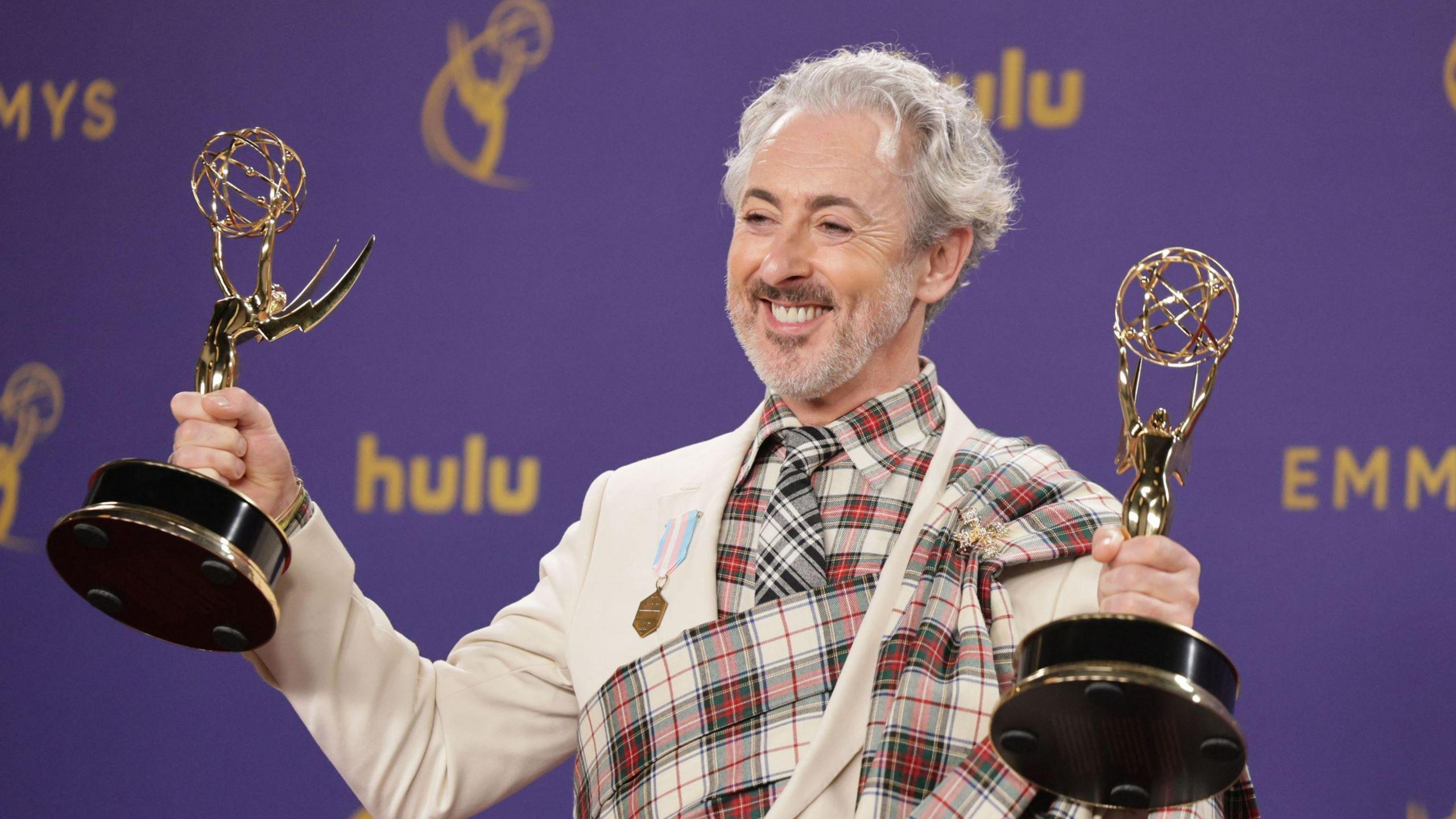 Alan Cumming celebrates the Traitors winning two Emmys in 2024 - he is grinning broadly while holding two Emmy awards up, one in each hand. He is wearing a suit with a tartan plaid design.