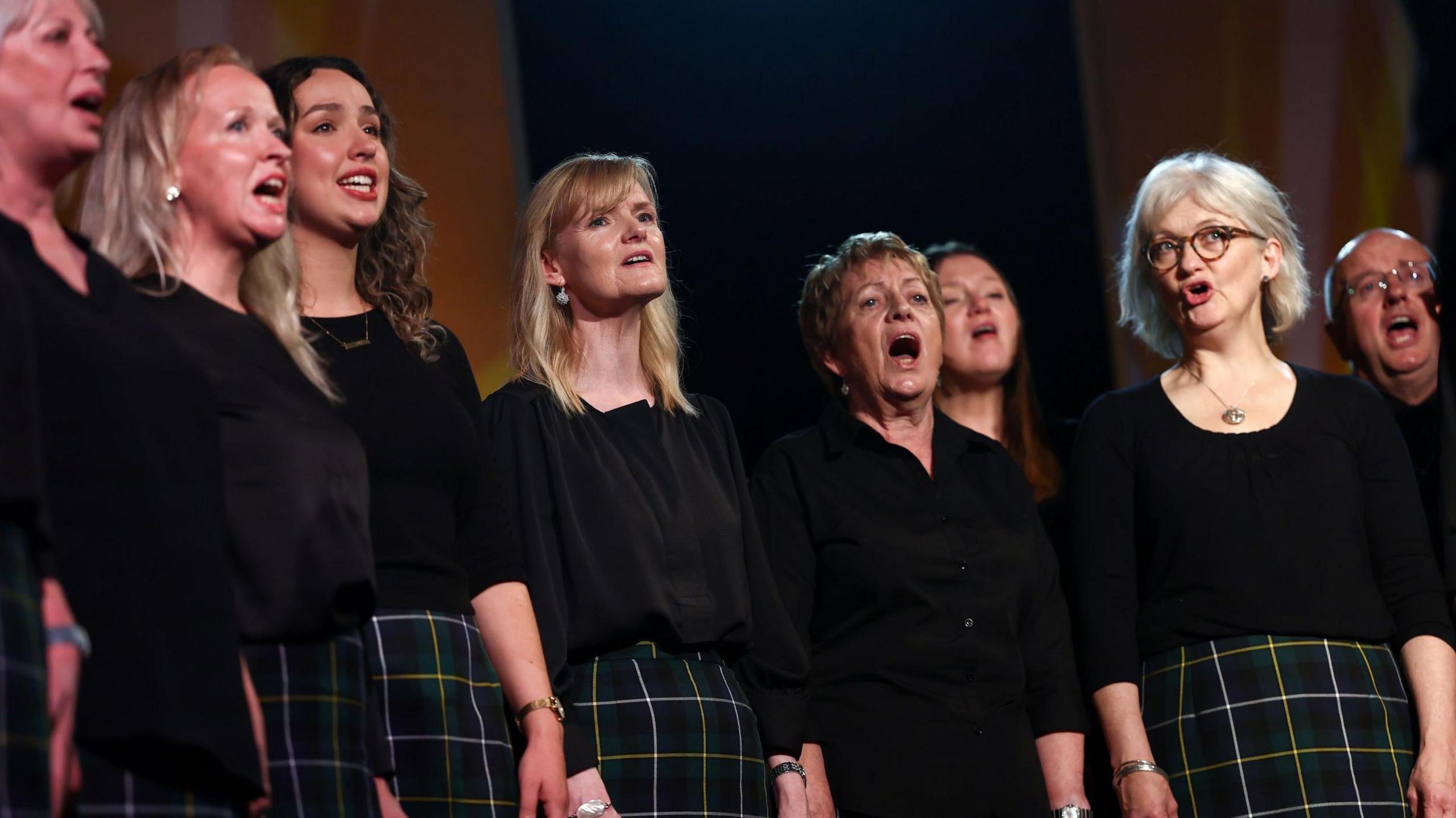 A Gaelic choir stands singing at the Mòd. The group is made up of female and male singers. They are wearing black tops and dark green tartan kilts.