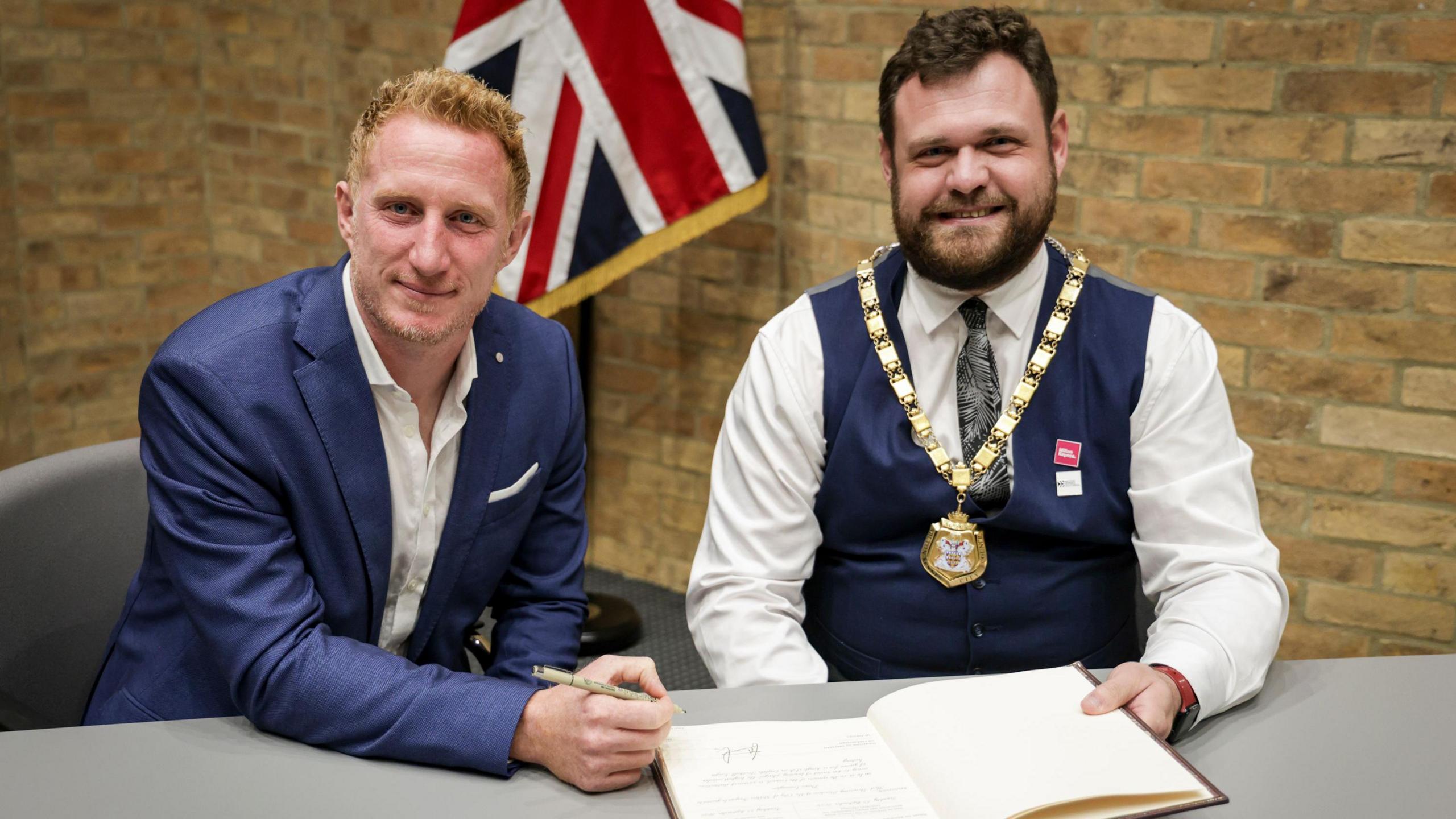 Two men sat at a table during a formal ceremony. Lewington wears a blue suit and holds a pen, while the mayor wears a vest, tie, and a ceremonial chain. An open book lies on the table, with a British flag in the background