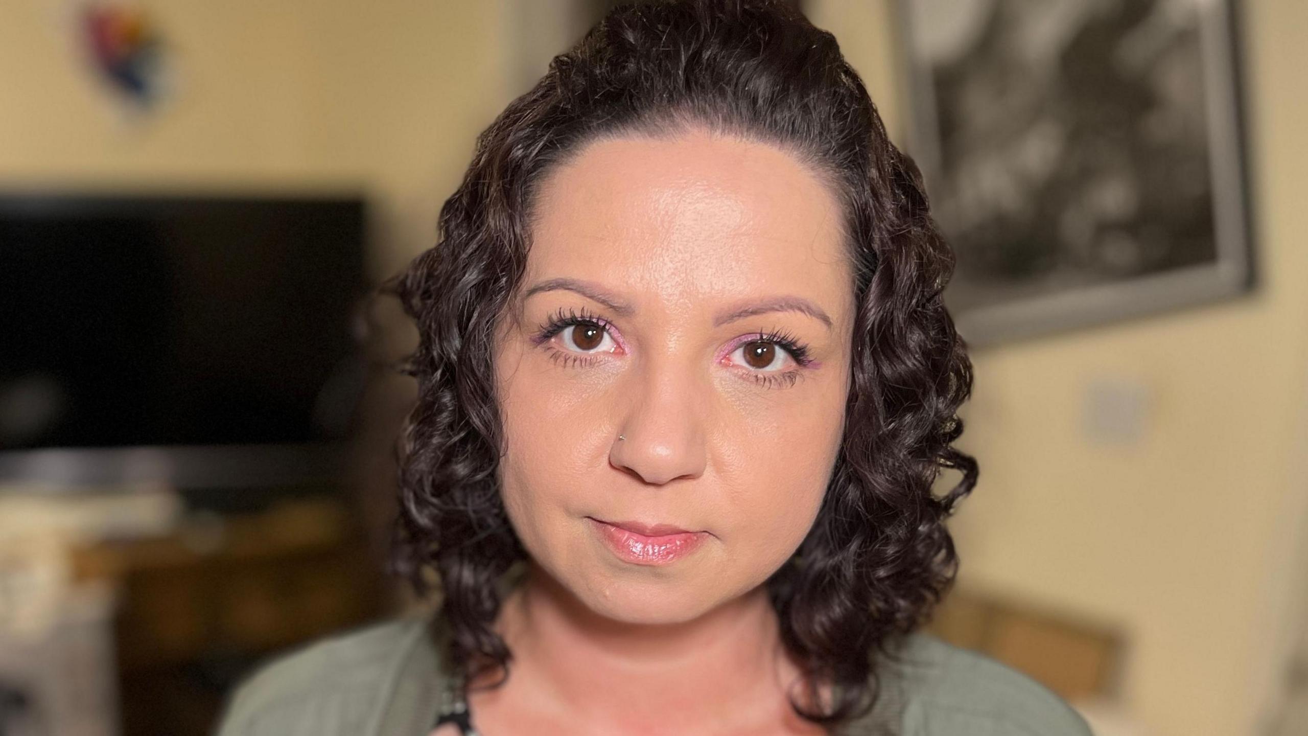 Eliza Suchocka inside her flat in Sycamore Field, Harlow. She is wearing a black top with silver studded detail, a green shirt and she has shoulder-length dark curly hair.