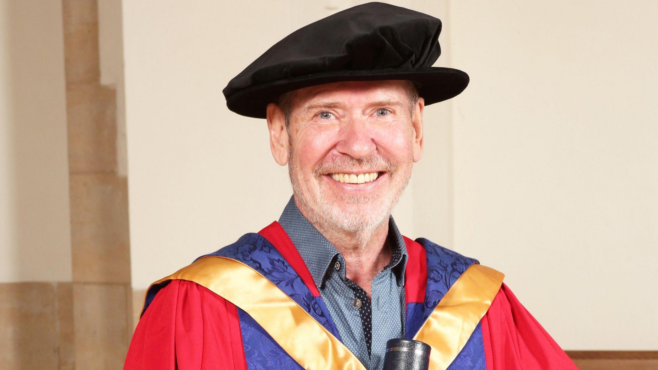 Steve Lillywhite, wearing a blue shirt underneath PhD robes and a hat, holds a degree scroll and smiles into the camera.