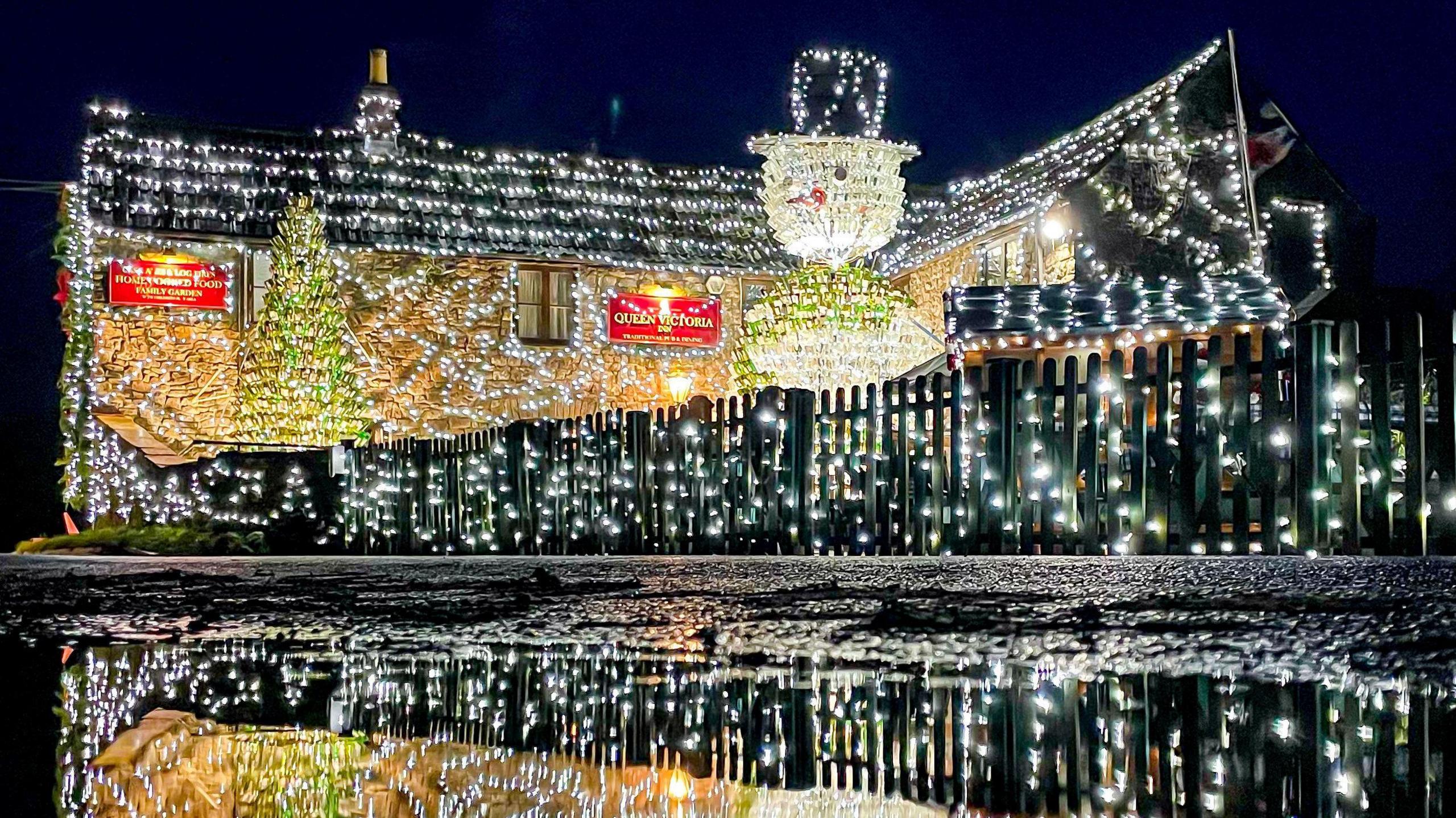 Queen Victoria pub in Priddy, Somerset. It is covered in white fairy lights. In front of the pub, there is a green Christmas tree display made out of empty beer bottles. It is almost as tall as the building. There is also a snowman made out of beer bottles. 