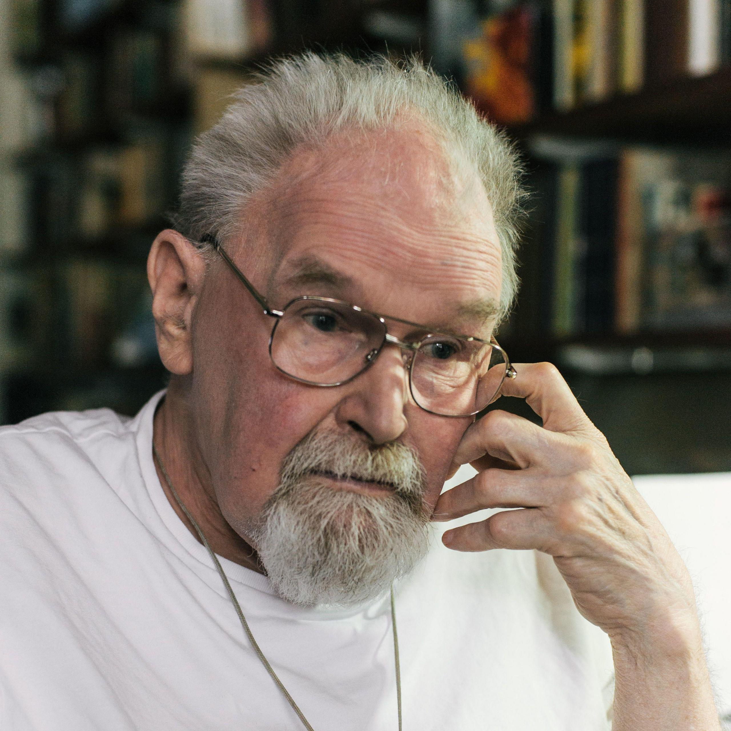 Alasdair Gray, a grey-haired man with a grey beard and moustache, stares solemnly to the side, his left hand at his cheek, wearing a plain white t-shirt and thin-rimmed glasses.