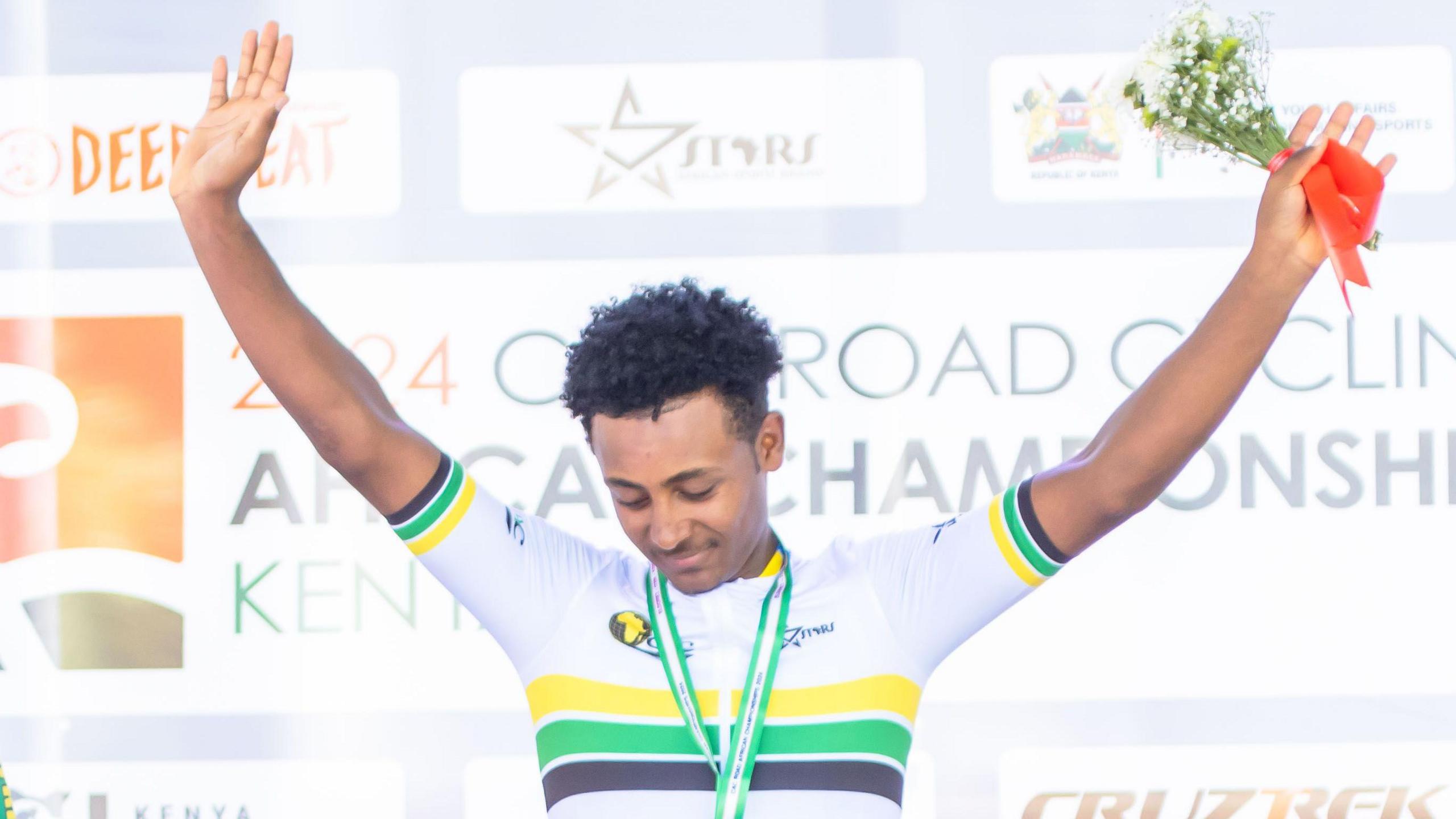 Keven Teklemariam puts both hands above his head in acclaim while holding a small bouquet of flowers while on the podium at the African Road Cycling Championships