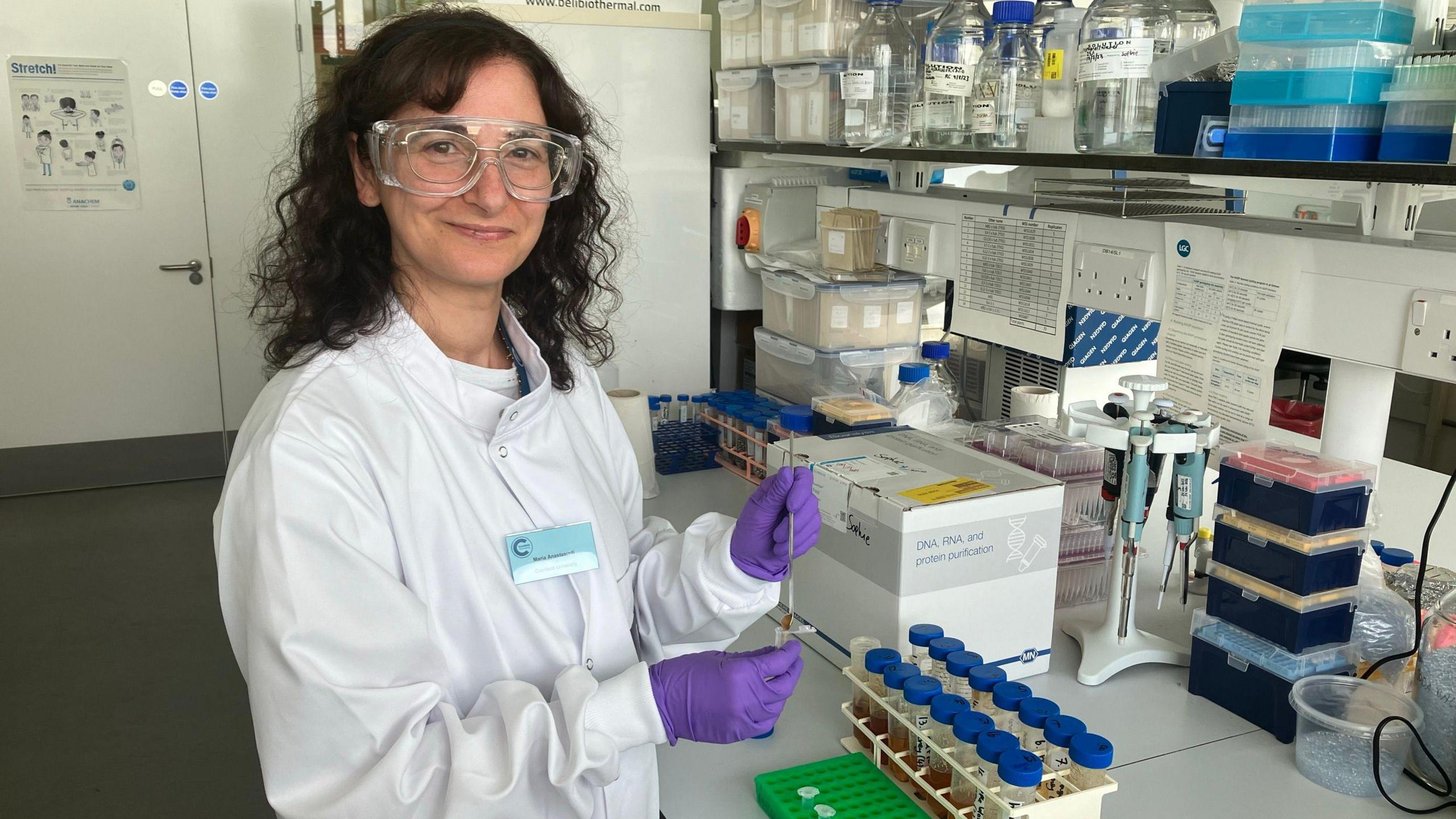 A picture of a dark haired woman, wearing safety goggles and a lab coat, in a laboratory, testing samples 