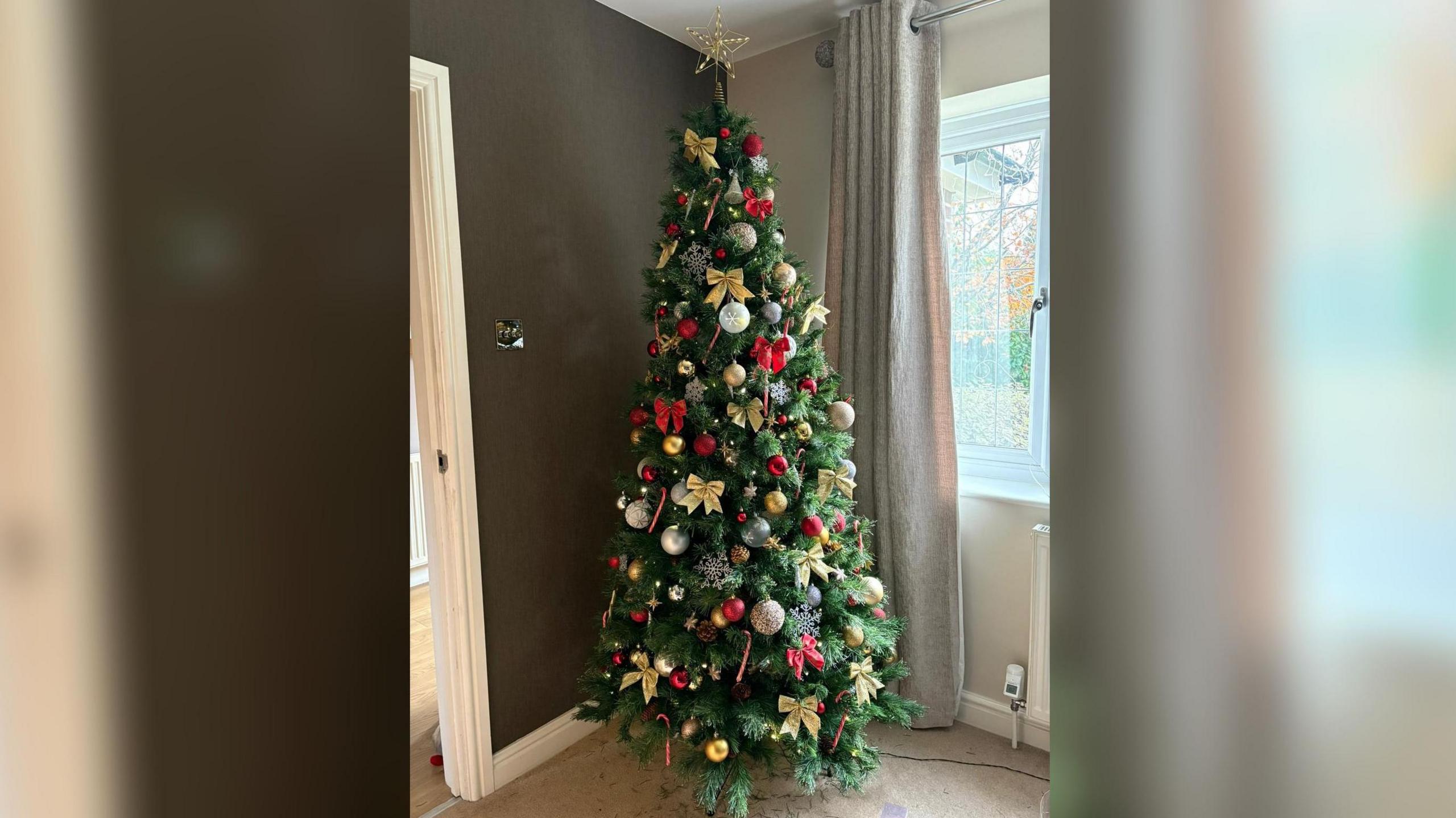 An artificial Christmas tree in the corner of a living room. It is decorated with gold bows, red and cold baubles and red striped candy canes.