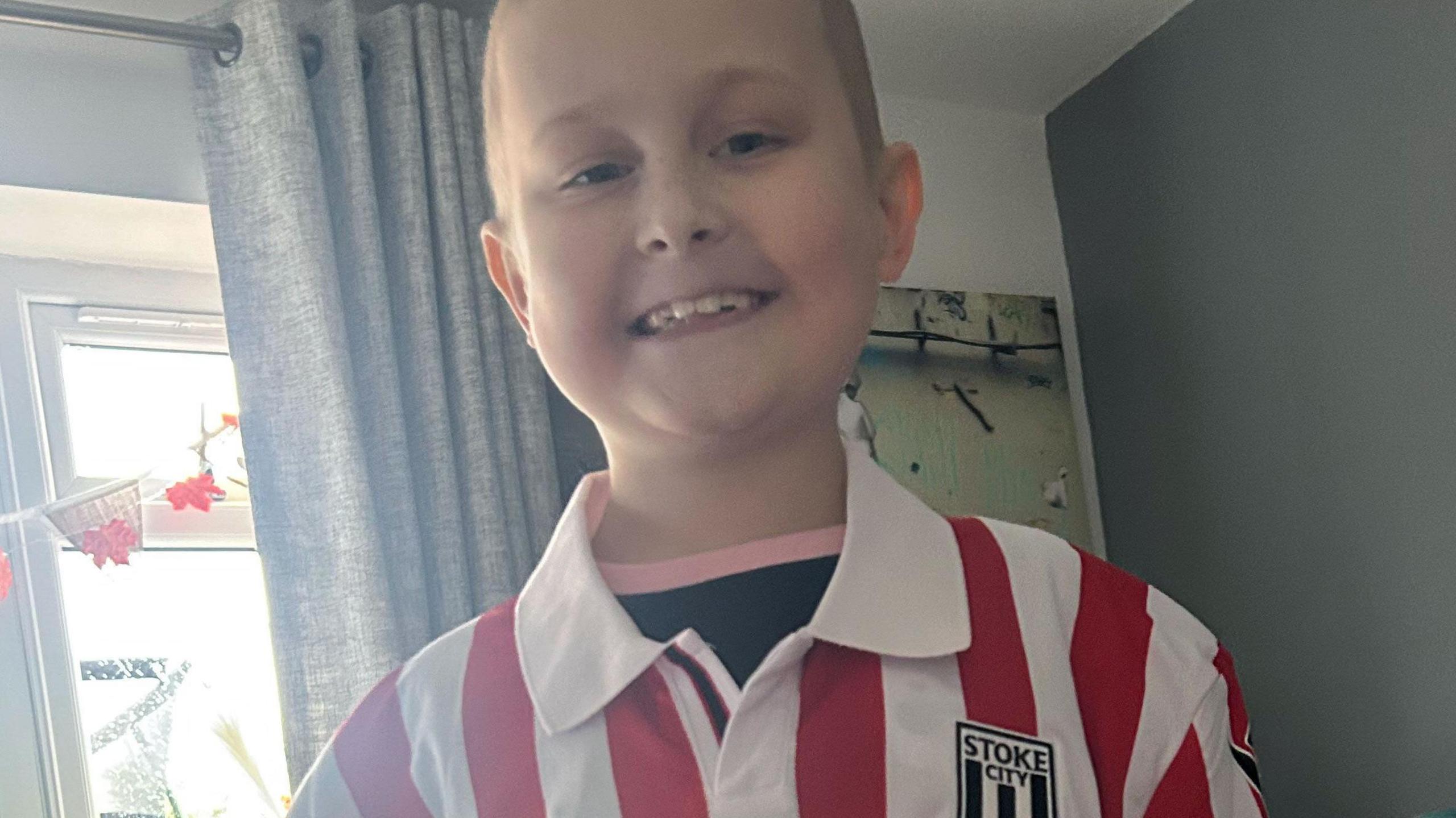 Harrison looking at the camera and smiling. He is wearing a red and white stipe Stoke City football shirt on top of a black t-shirt with a pink collar. Behind him are curtains and window decorations. 