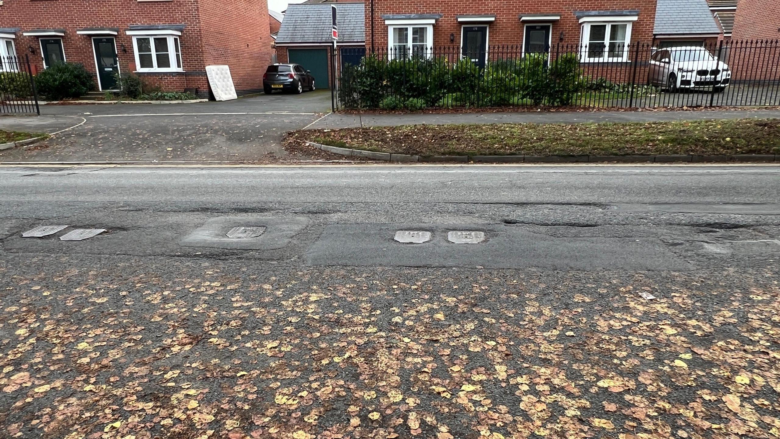 Image shows a tarmac road with pot holes filled in with cement, leaving the road uneven. Two read brick houses can be seen on the far side of the road. 