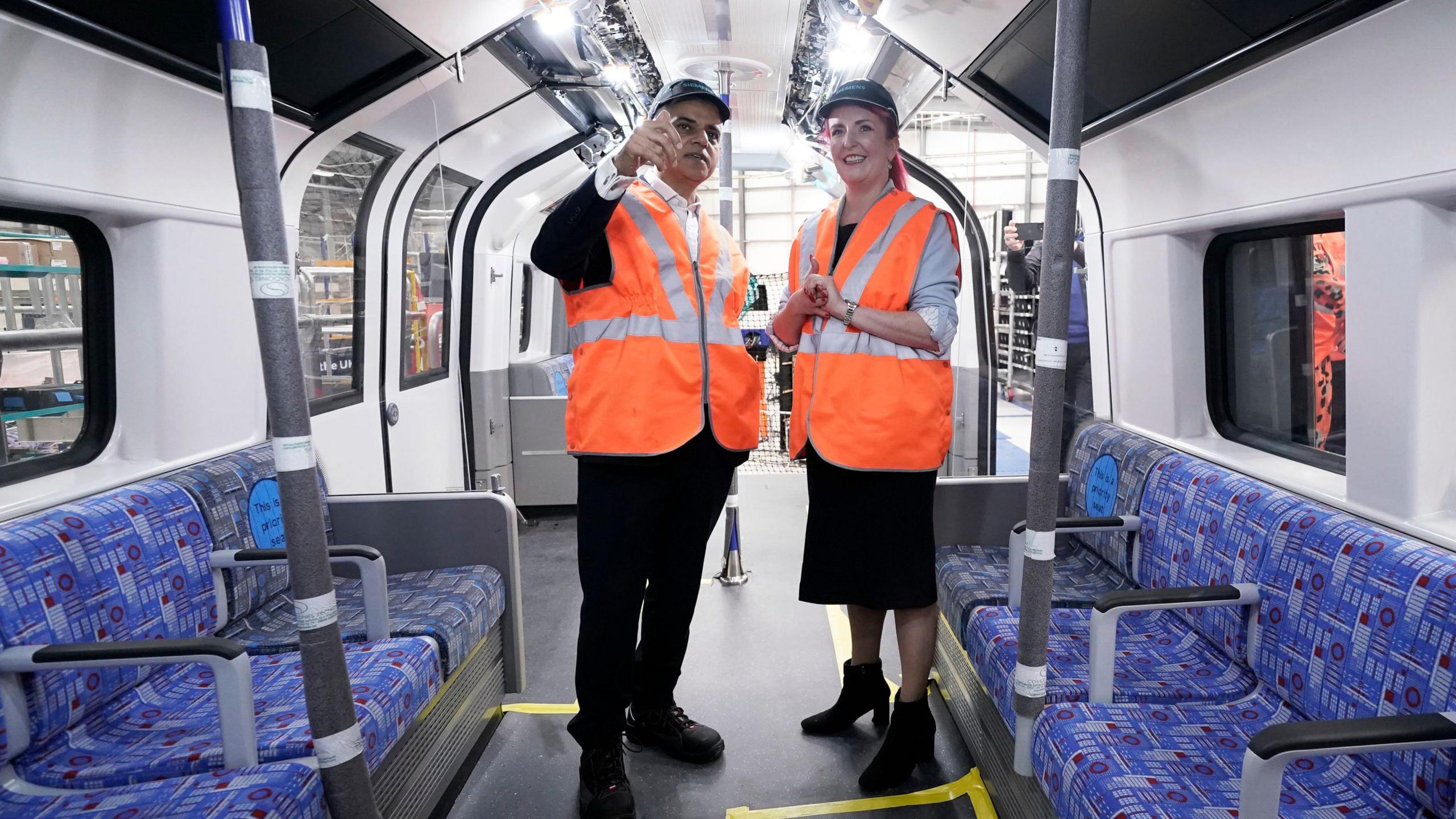 Sadiq and Louise are standing inside an unfinished tube train carriage. Sadiq is on the left and he is wearing an orange high-vis vest and a navy cap with Siemens on it. He has his right-hand raised and is looking forward. Louise is standing on the right at an angle facing Sadiq. She is also wearing an orange high-vis vest and a navy cap with Siemens on it. Both her hands are clasped together at the waist