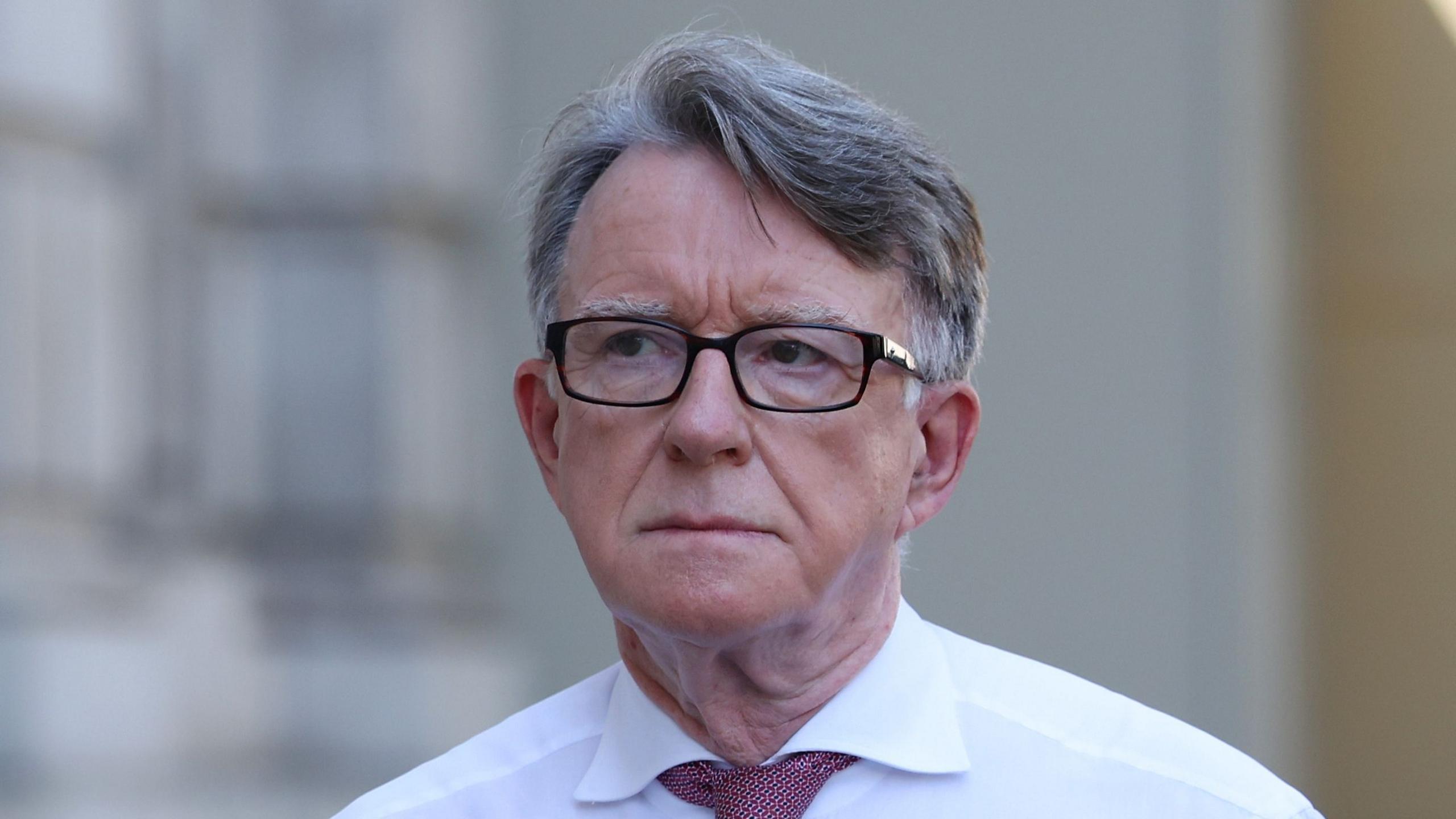 Peter Mandelson is wearing a pair of glasses and a white shirt with a red tie.